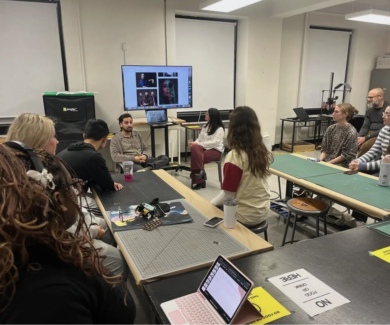 A group of people attending a photo workshop in a classroom with tables, a large monitor displaying photos, and various photography equipment.
