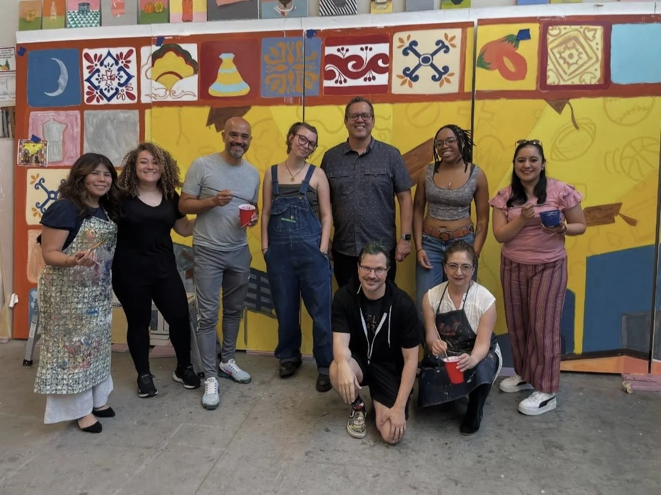 A group of eleven people, smiling and posing for a photo in front of a colorful mural with various icons. Some are holding paint supplies and red cups, indicating an art or community event.