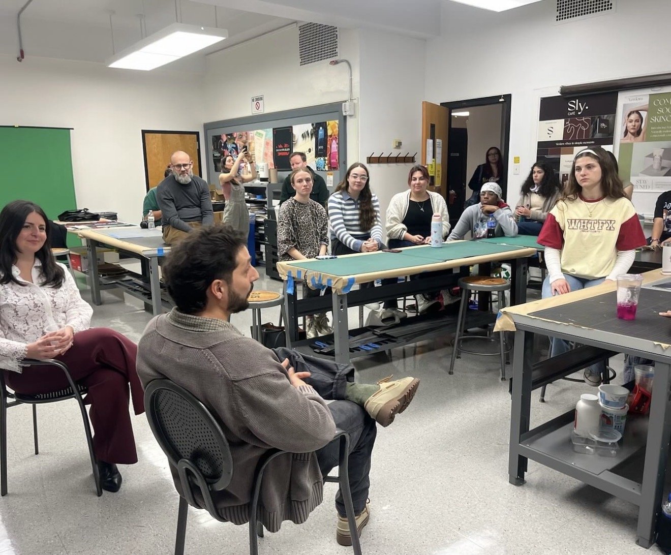 A group of people in a classroom or workshop setting, seated around tables and listening to a speaker or presenter. Some individuals are taking notes, others are sitting attentively, and a few are standing near the back.