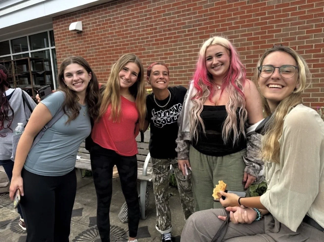 Group of five young women smiling and posing outdoors in front of a brick wall, with one woman holding a piece of slice of pizza.