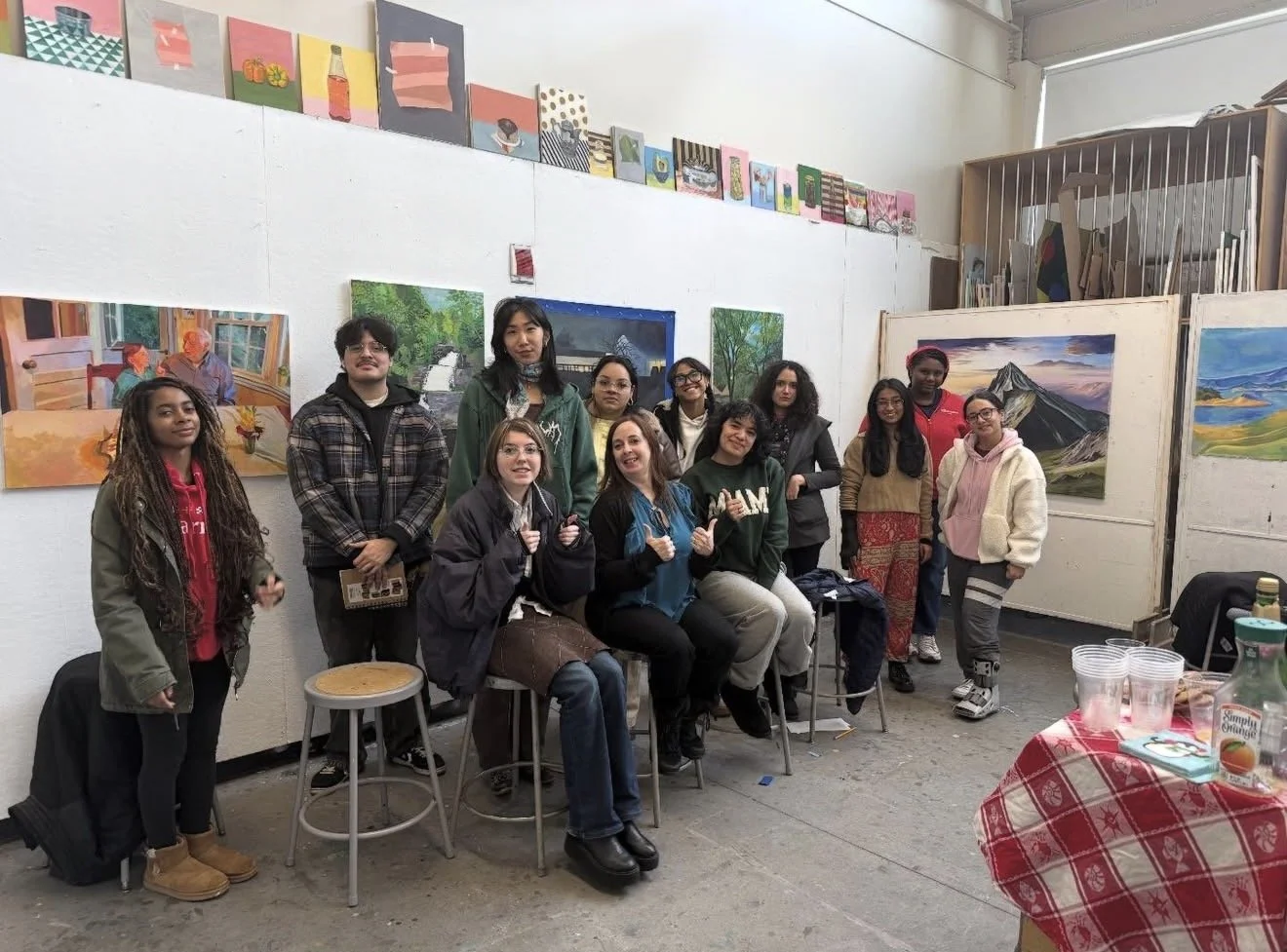 Group of people in an art gallery with landscape paintings hanging on the walls, some seated on stools, some standing, a table with drinks and snacks on the right.