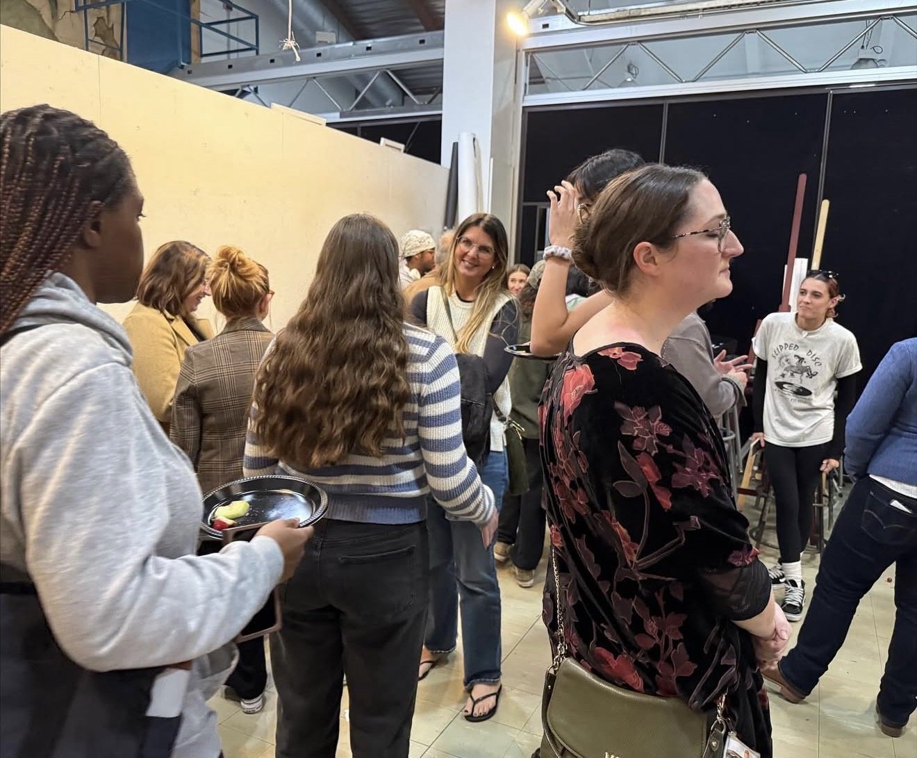 Group of women standing and mingling in an indoor space, some talking and others waiting, with some holding small plates of food.