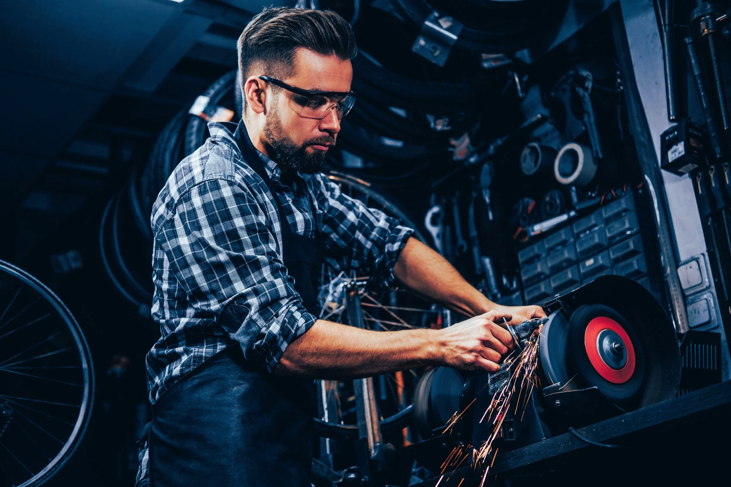 A man wearing safety glasses and a plaid shirt works in a manufacturing facility, using a grinding tool that produces sparks.