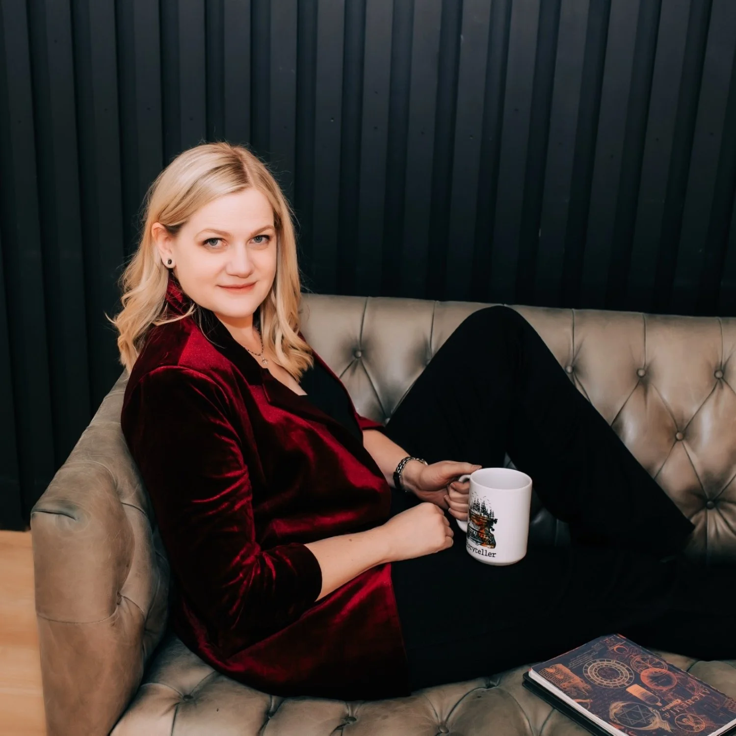 A woman with blonde hair sitting on a beige tufted leather couch, holding a white mug with a Christmas tree design, in front of a dark paneled wall, and a notebook on the couch.