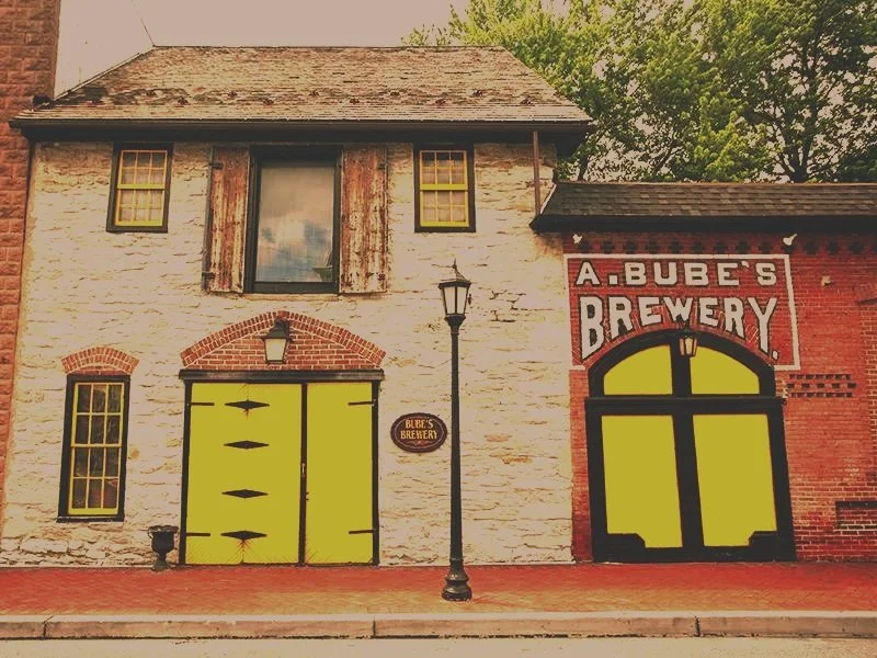 Exterior of a building with a sign reading 'A. Bube's Brewery', painted yellow doors, brick and white painted brick walls, and a lamp post in front.