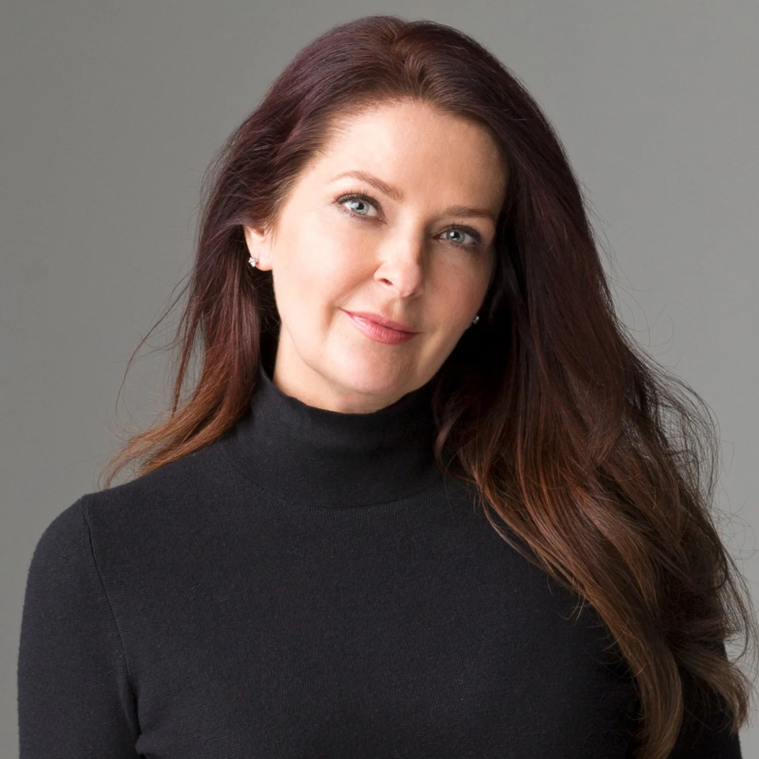 A woman with long auburn hair wearing a black turtleneck, smiling softly against a plain gray background.