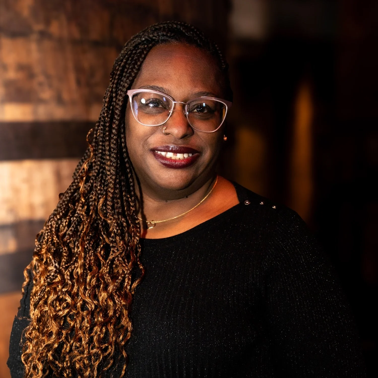 A woman with dark skin, long curly hair, wearing glasses, a nose ring, small earrings, and a black top, smiling at the camera in a dimly lit indoor setting.