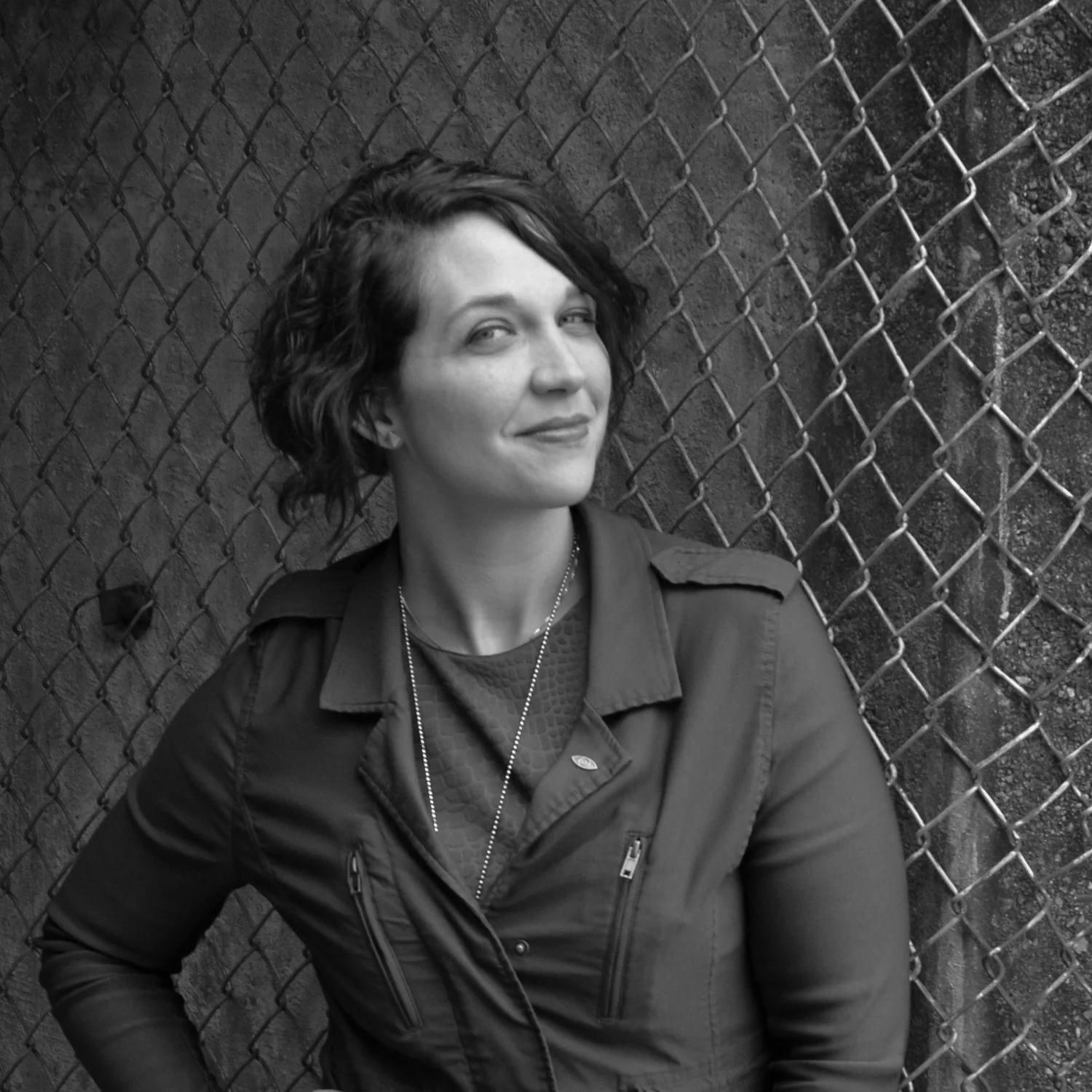A woman with short wavy hair smiling and leaning against a chain-link fence.