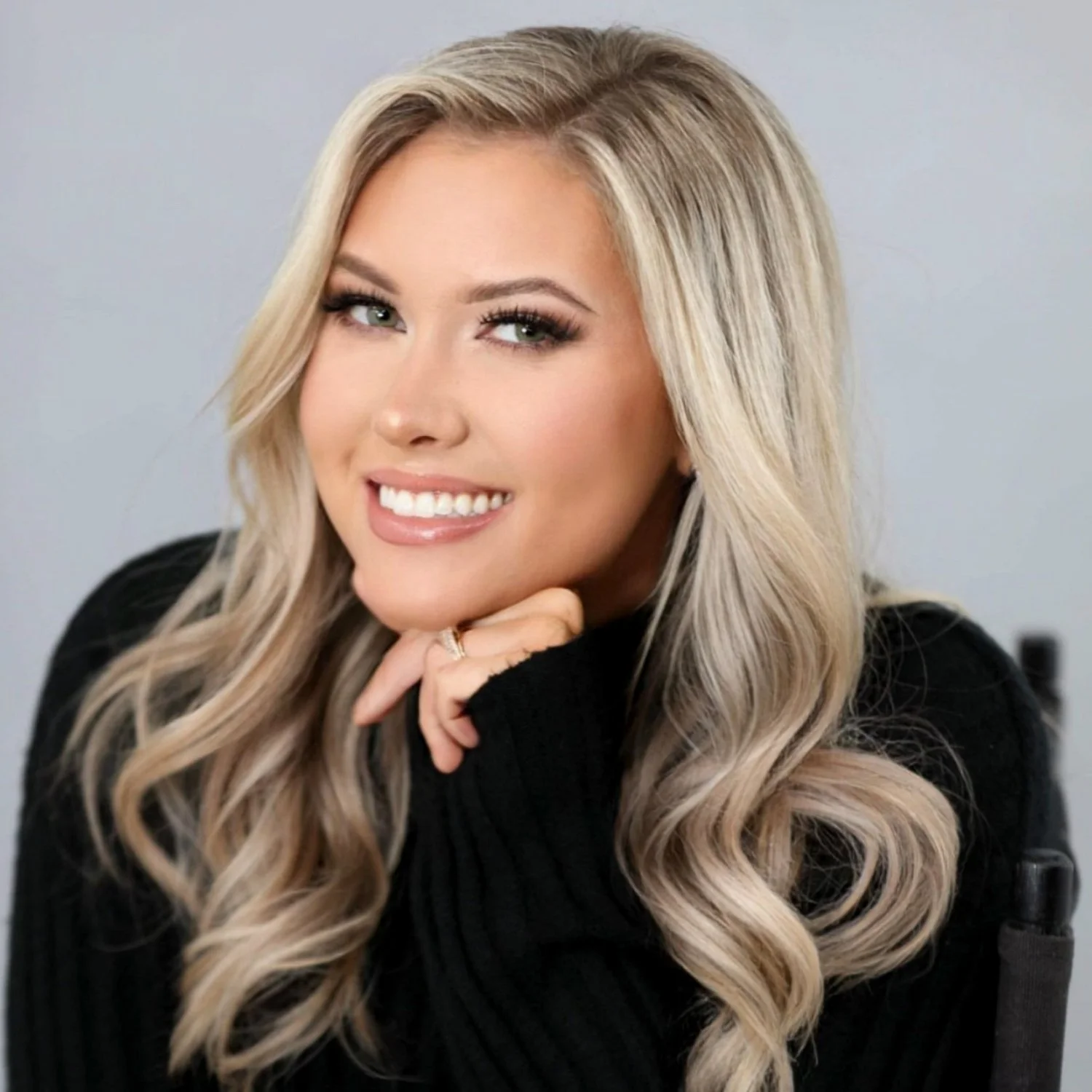 Close-up portrait of a young blonde woman with wavy hair, smiling, wearing a black sweater, resting her chin on her hand, in a professional setting.