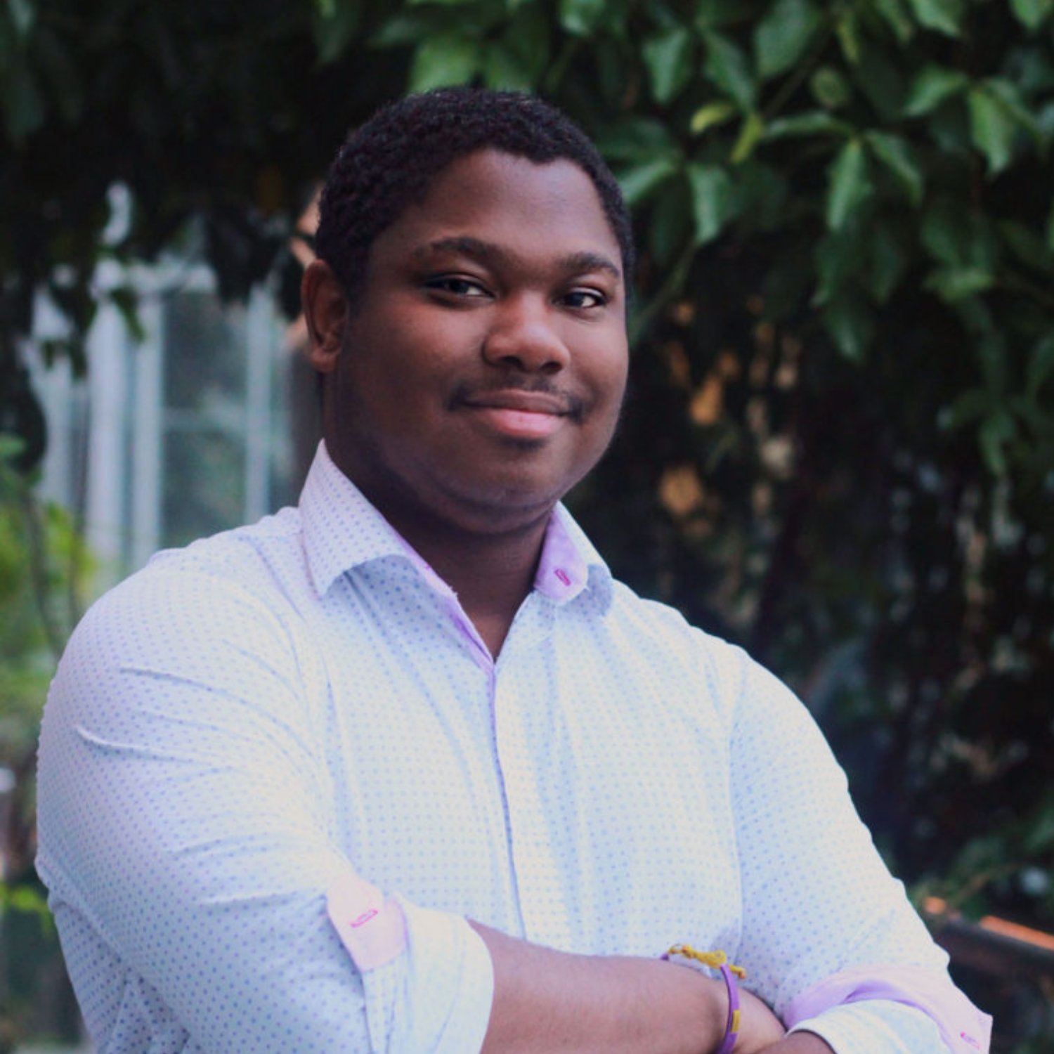 Young man with crossed arms standing outdoors in front of green foliage, smiling at the camera.
