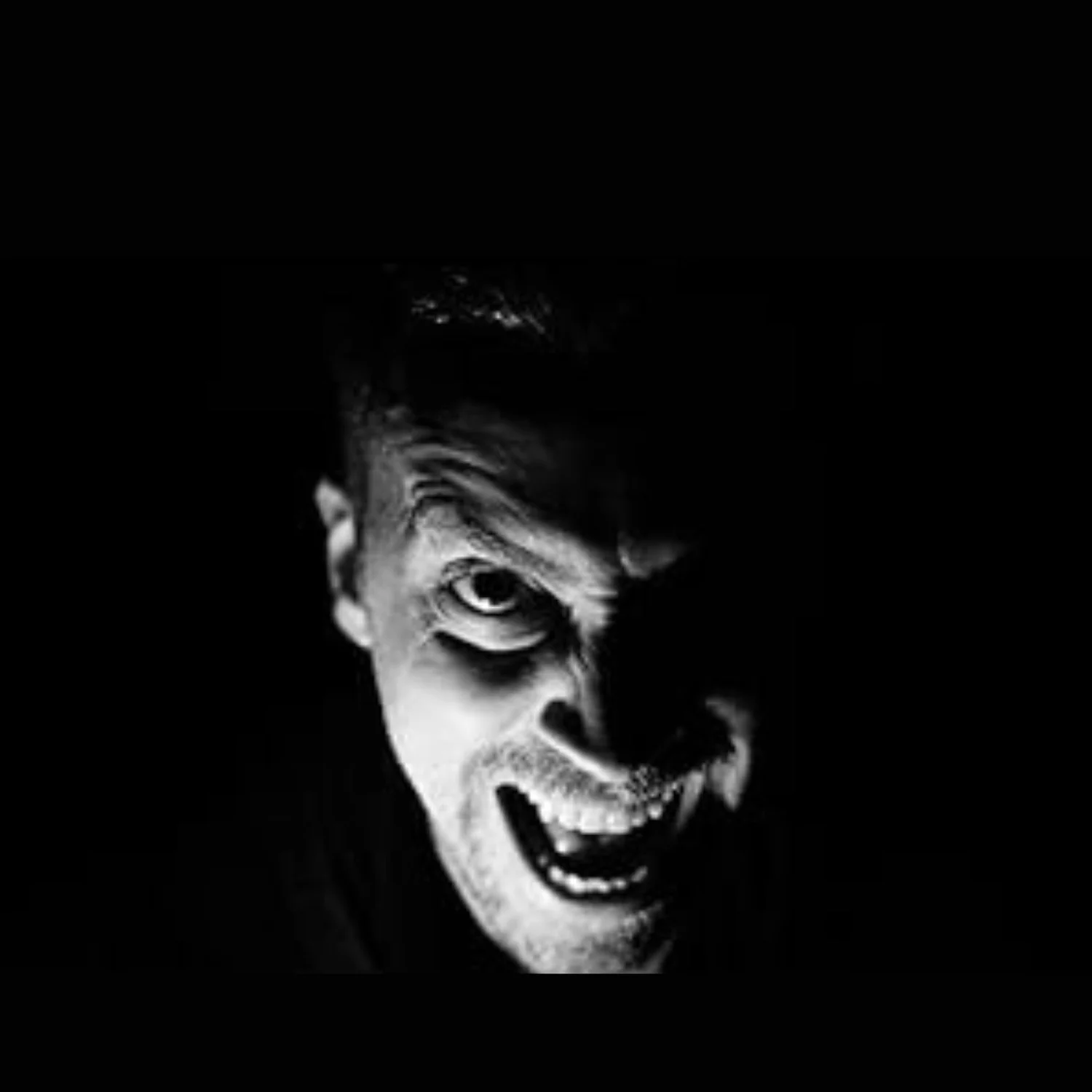 Black and white close-up of a man with a manic expression, one eye wide open, mouth open showing teeth, with shadows emphasizing facial features.