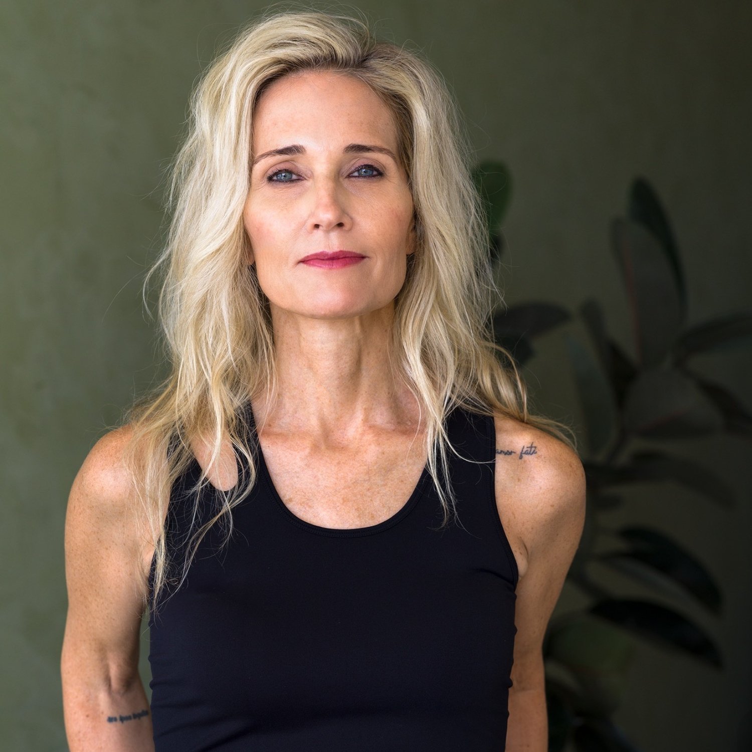 Portrait of a mature woman with blonde hair, wearing a black sleeveless top, standing in front of a green wall with a plant in the background.