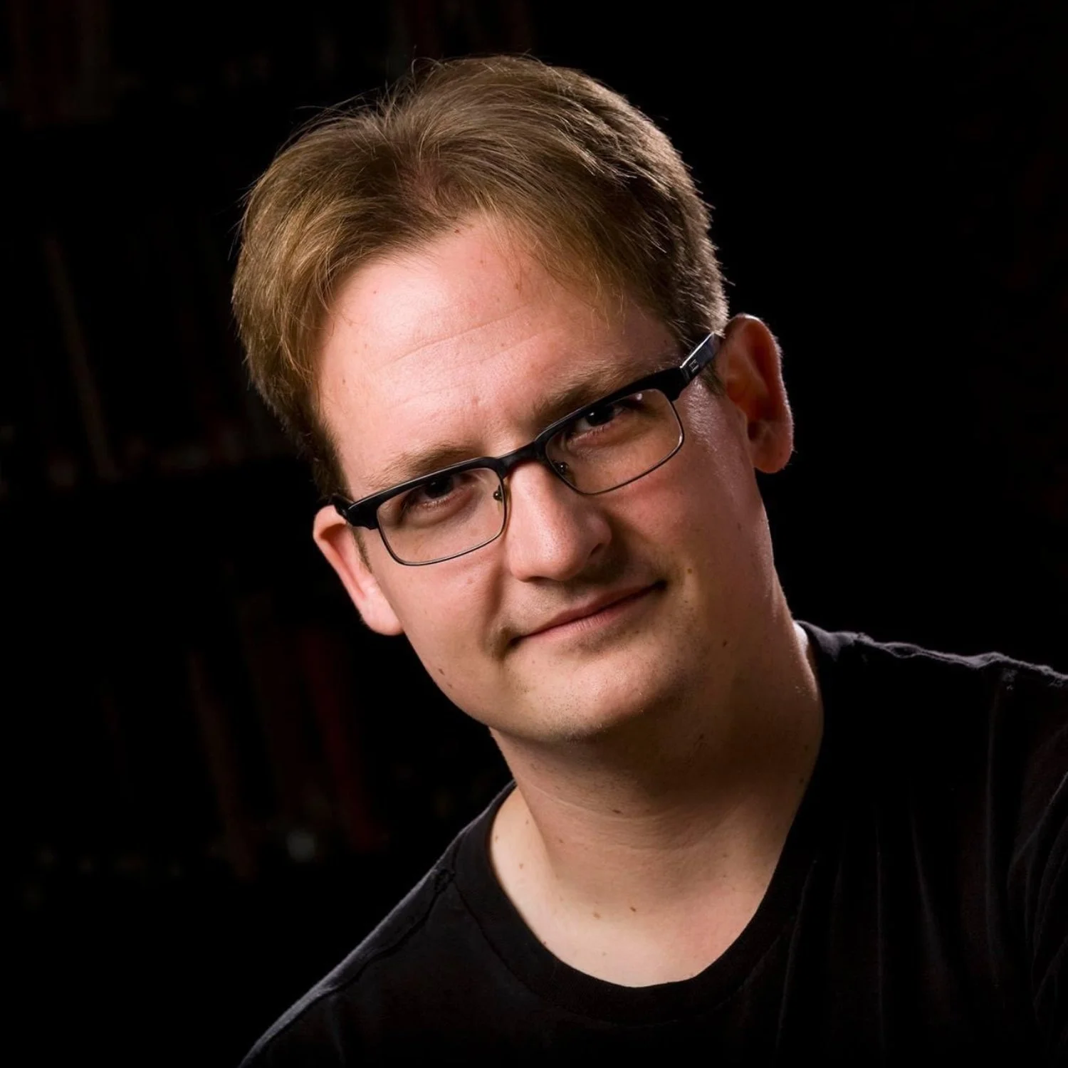 A young man with light brown hair and glasses, wearing a black shirt, posing against a dark background.