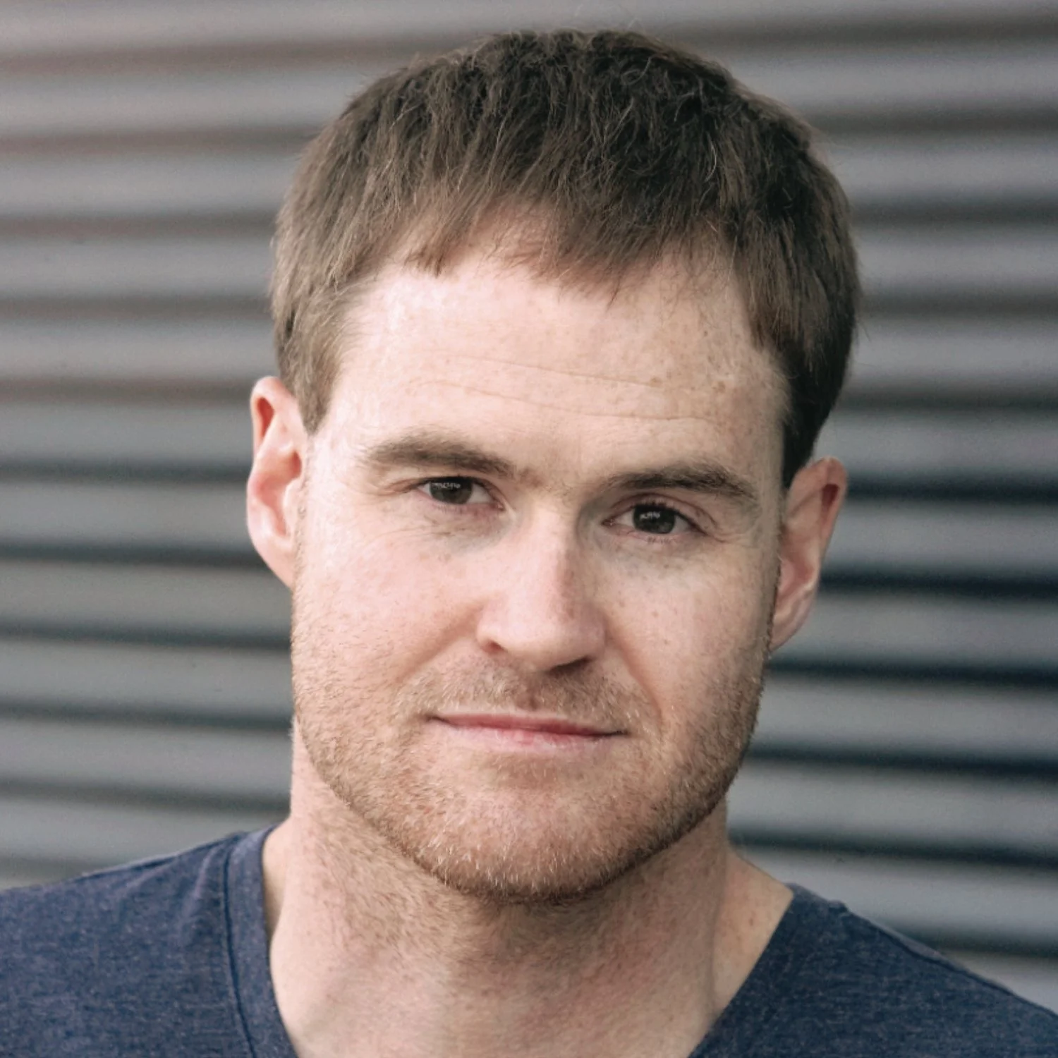 Close-up portrait of a young man with short brown hair and light skin, wearing a dark T-shirt, standing outdoors in front of a metal siding background.
