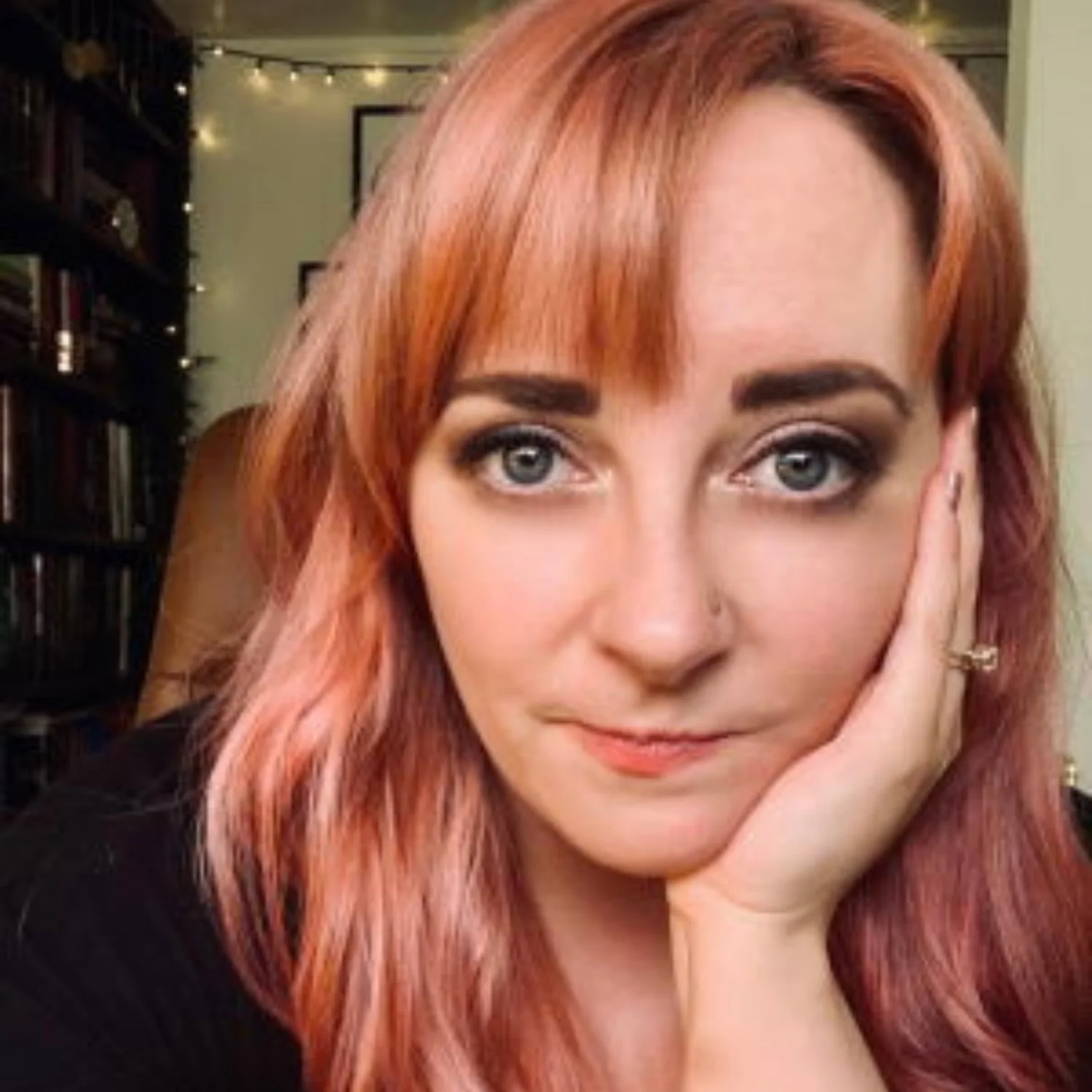 A woman with reddish-blonde hair and light blue eyes rests her face on her hand, seated in a room with bookshelves and warm lighting.