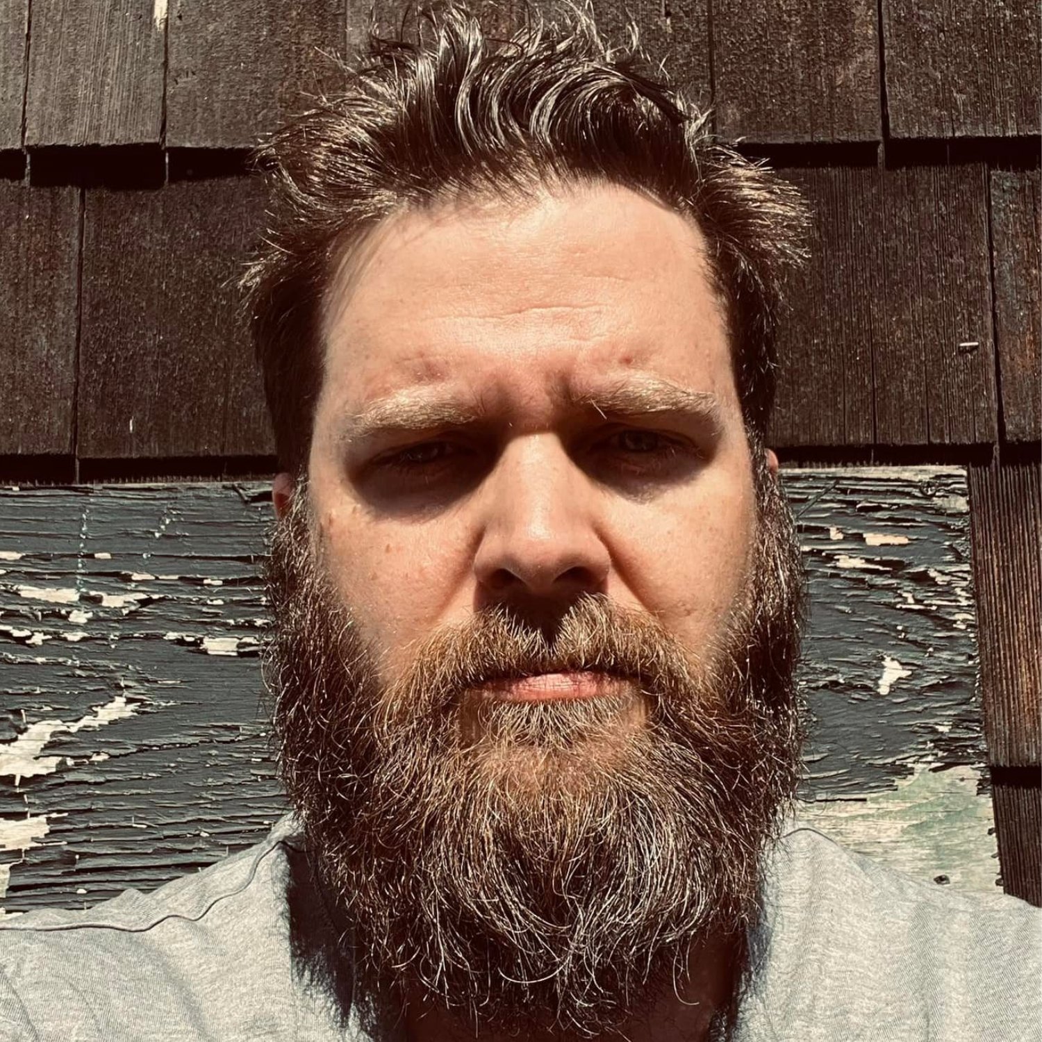 A close-up of a man with a beard and messy hair, looking into the camera, with a weathered wooden wall in the background.