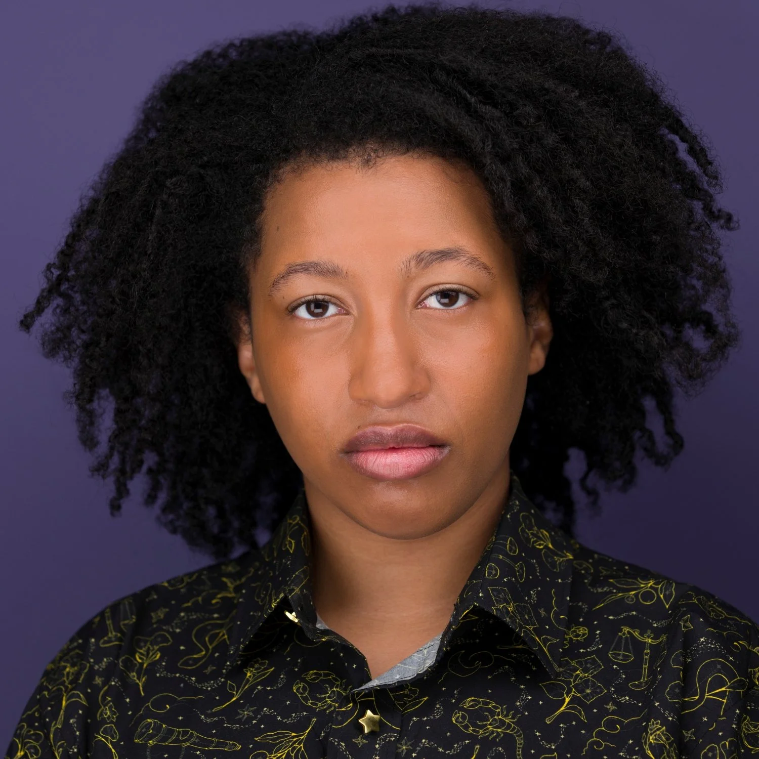 Portrait of a woman with natural curly black hair, wearing a black shirt with yellow line drawings of various symbols, against a dark purple background.