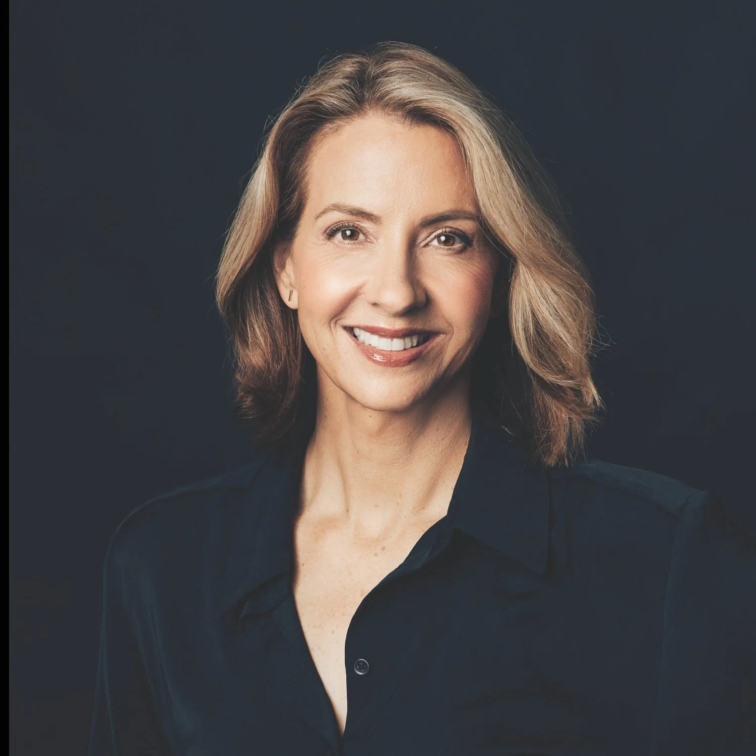 Portrait of a smiling woman with shoulder-length wavy hair, wearing a dark shirt, against a dark background.