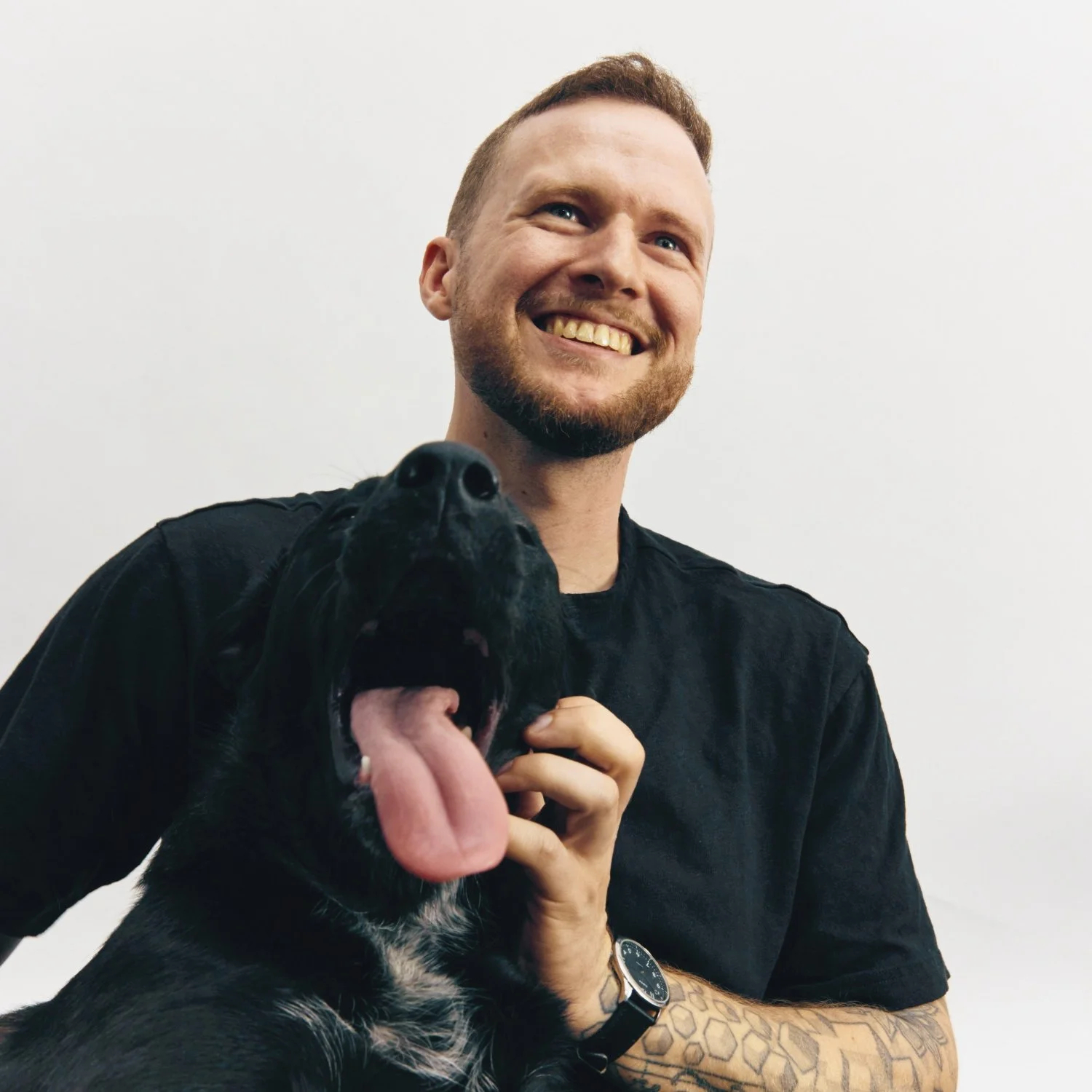 A smiling man with a beard and tattooed arm holding a black dog with its tongue out against a plain white background.
