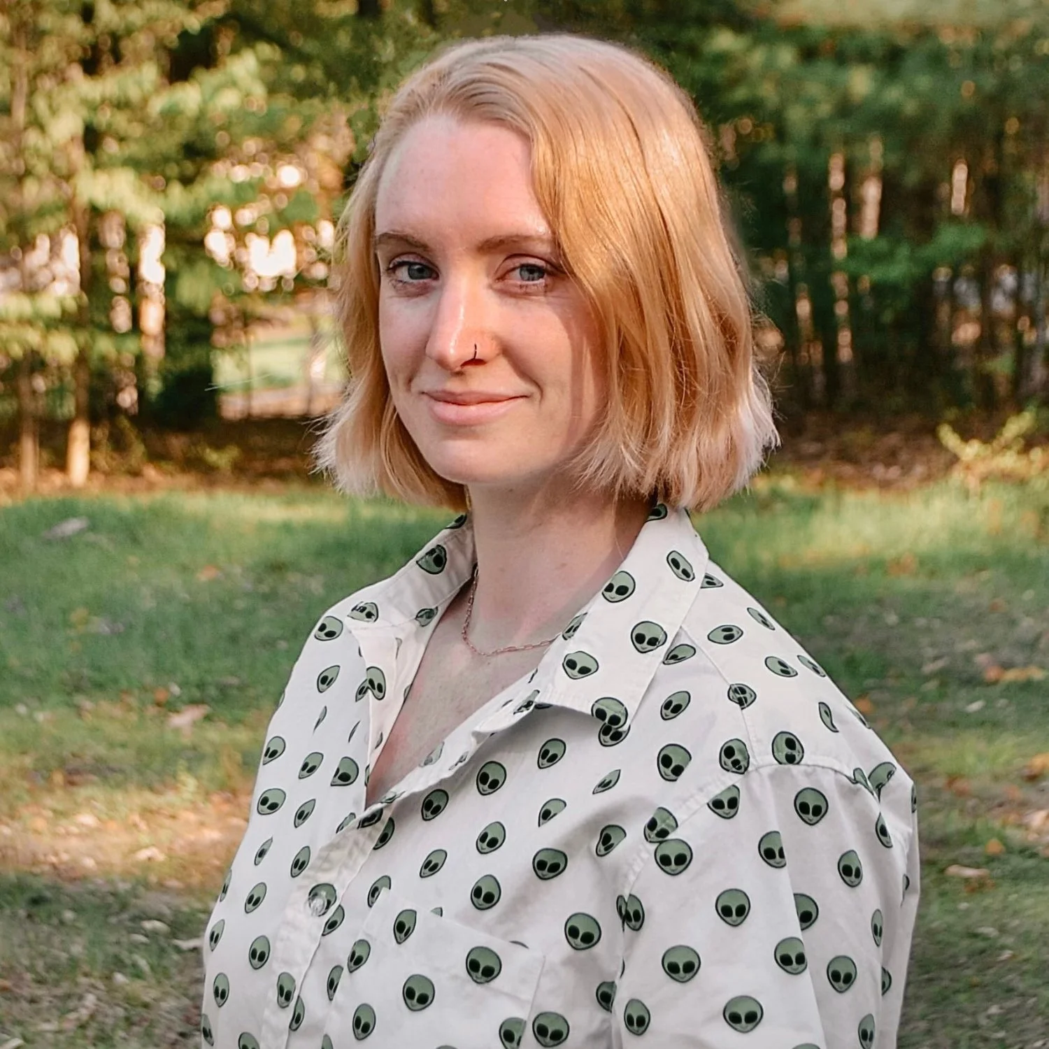 A woman with shoulder-length strawberry blonde hair and blue eyes, wearing a white shirt with a pattern of green alien faces, stands outdoors in a park with trees and grass in the background.