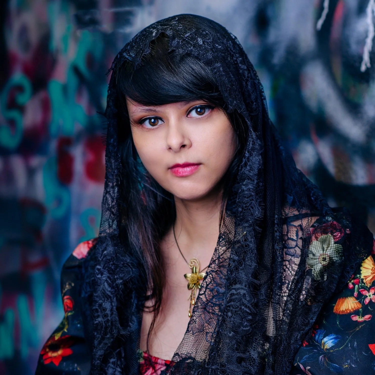 A woman wearing a black lace head covering and floral dress stands in front of a graffiti-covered wall.
