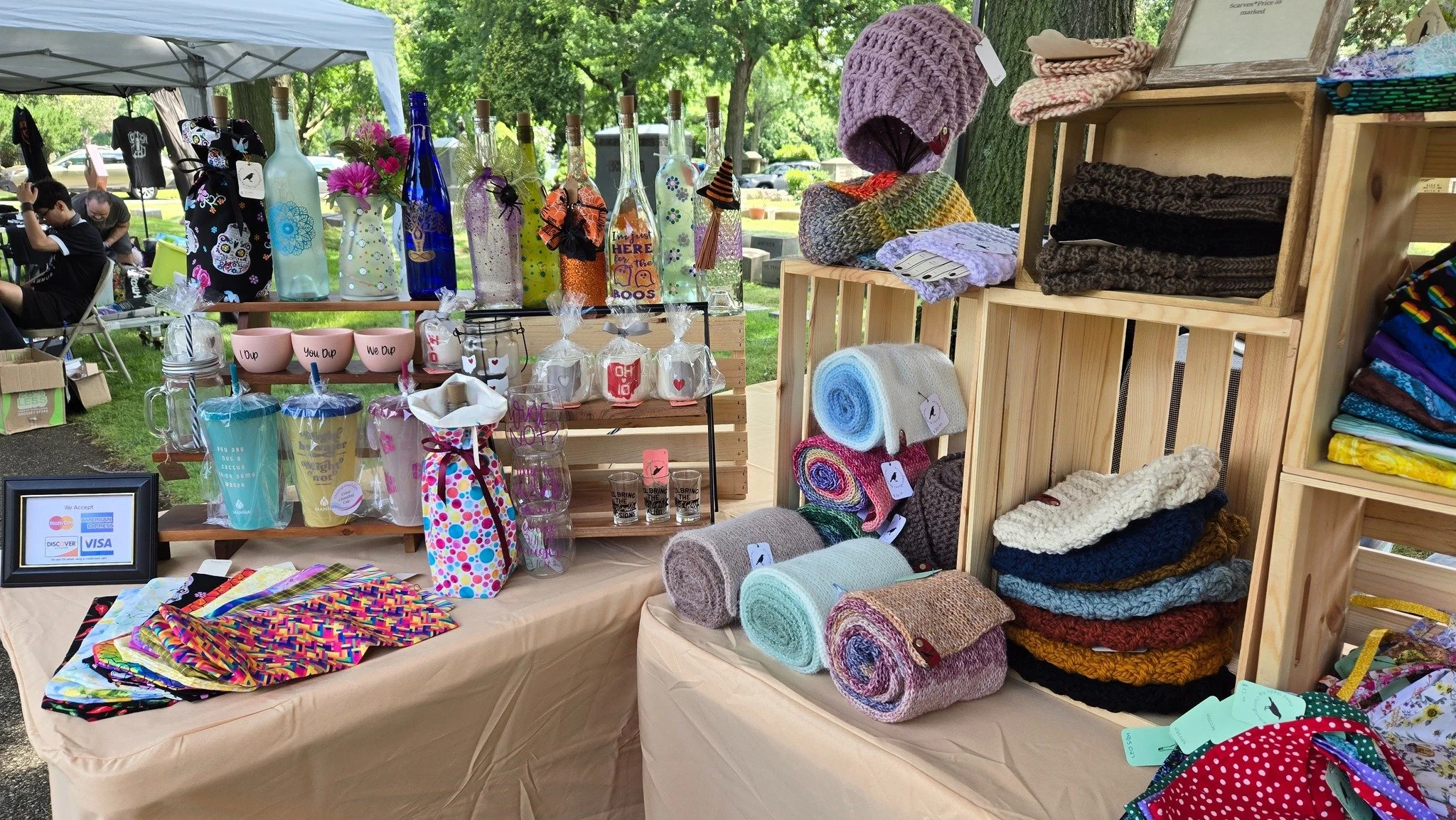 A display of handmade crafts and clothing at an outdoor market stall. The stall features knitted hats, towels, and fabric items, with bottles decorated for sale, under a canopy with trees in the background.