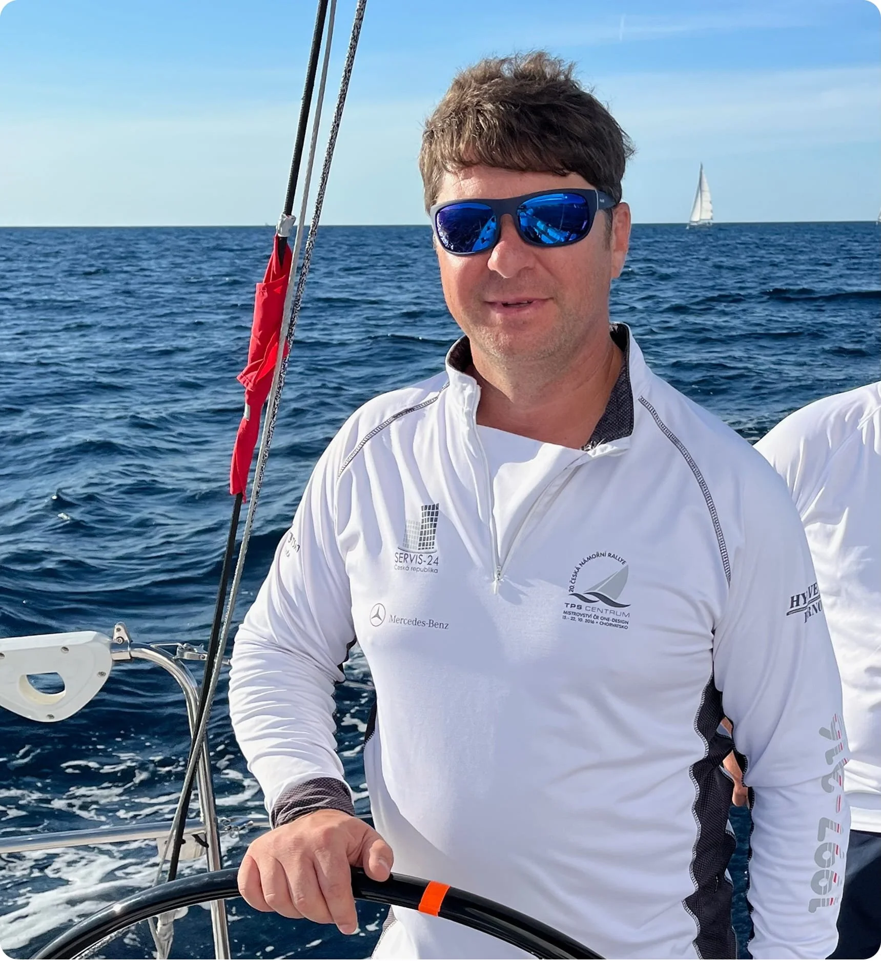 A man with brown hair and blue sunglasses taking a selfie on a boat, with the sea and a distant sailboat in the background, wearing a white sports jacket with logos.