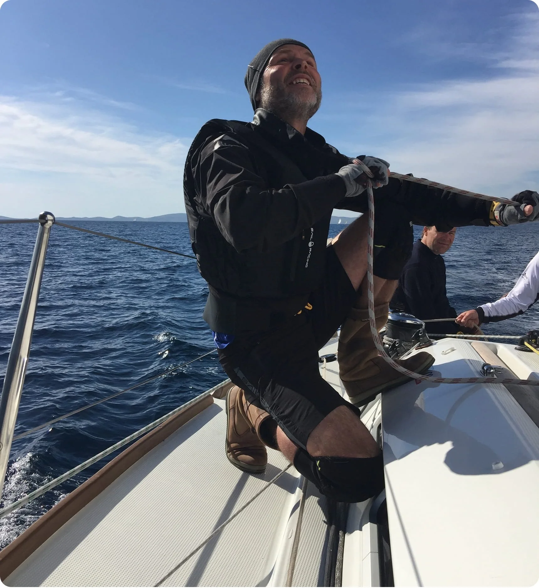 A man on a boat holding a rope, sailing in the ocean with a clear blue sky and distant islands in the background.