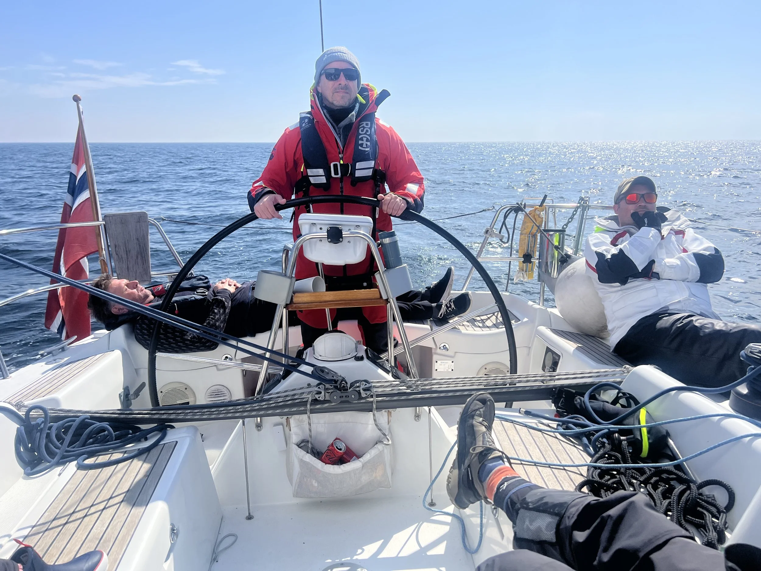 Two men on a sailboat in open water with clear skies. One man is at the helm wearing sunglasses and a gray beanie, while the other man is reclining on the deck wearing sunglasses and a white jacket. The boat has various ropes and equipment, and a red flag is visible on the stern.