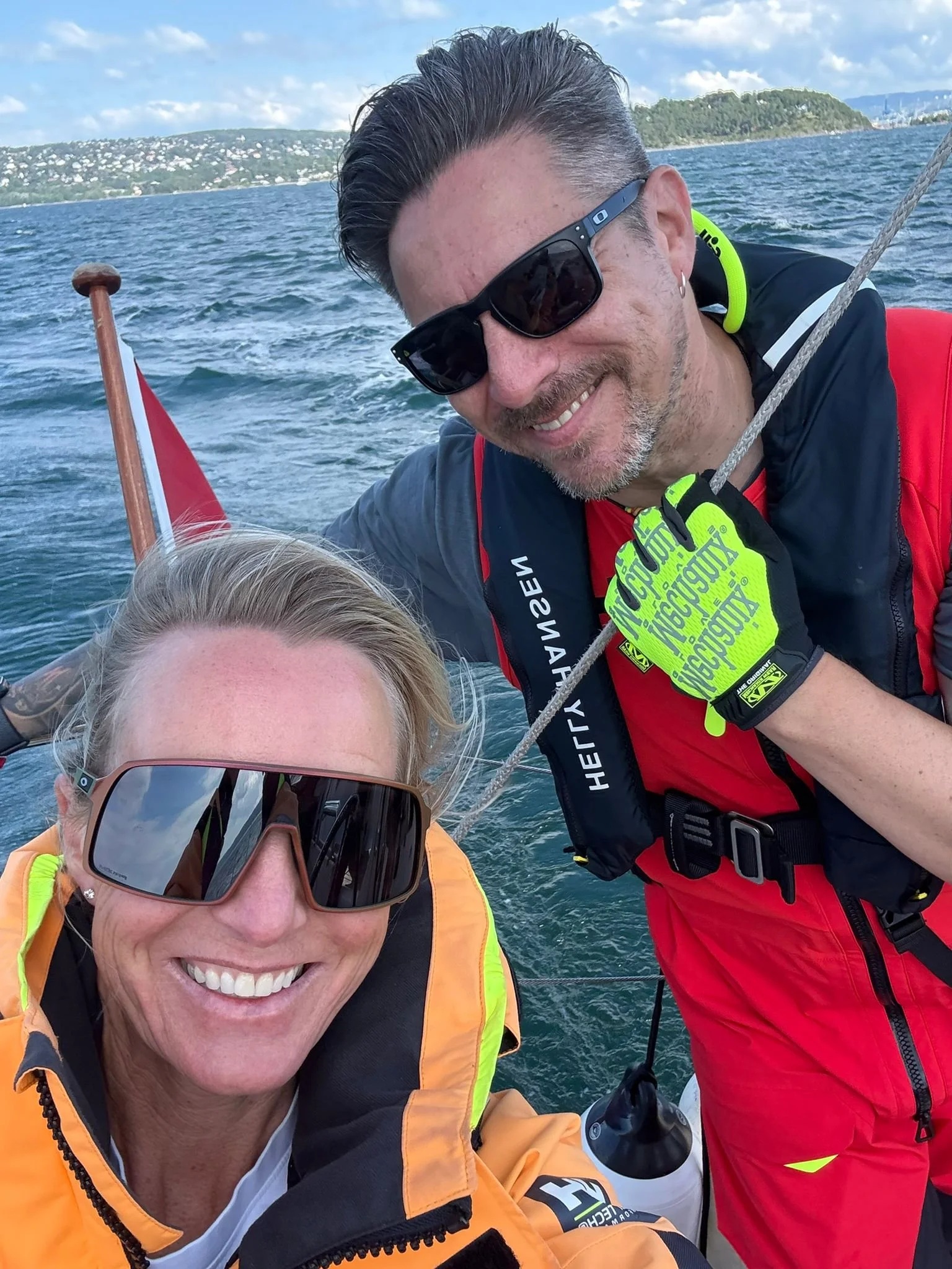 A smiling woman wearing sunglasses and an orange jacket, and a smiling man with sunglasses and a dark jacket, posing on a boat with water, a distant shoreline, and partly cloudy sky in the background.