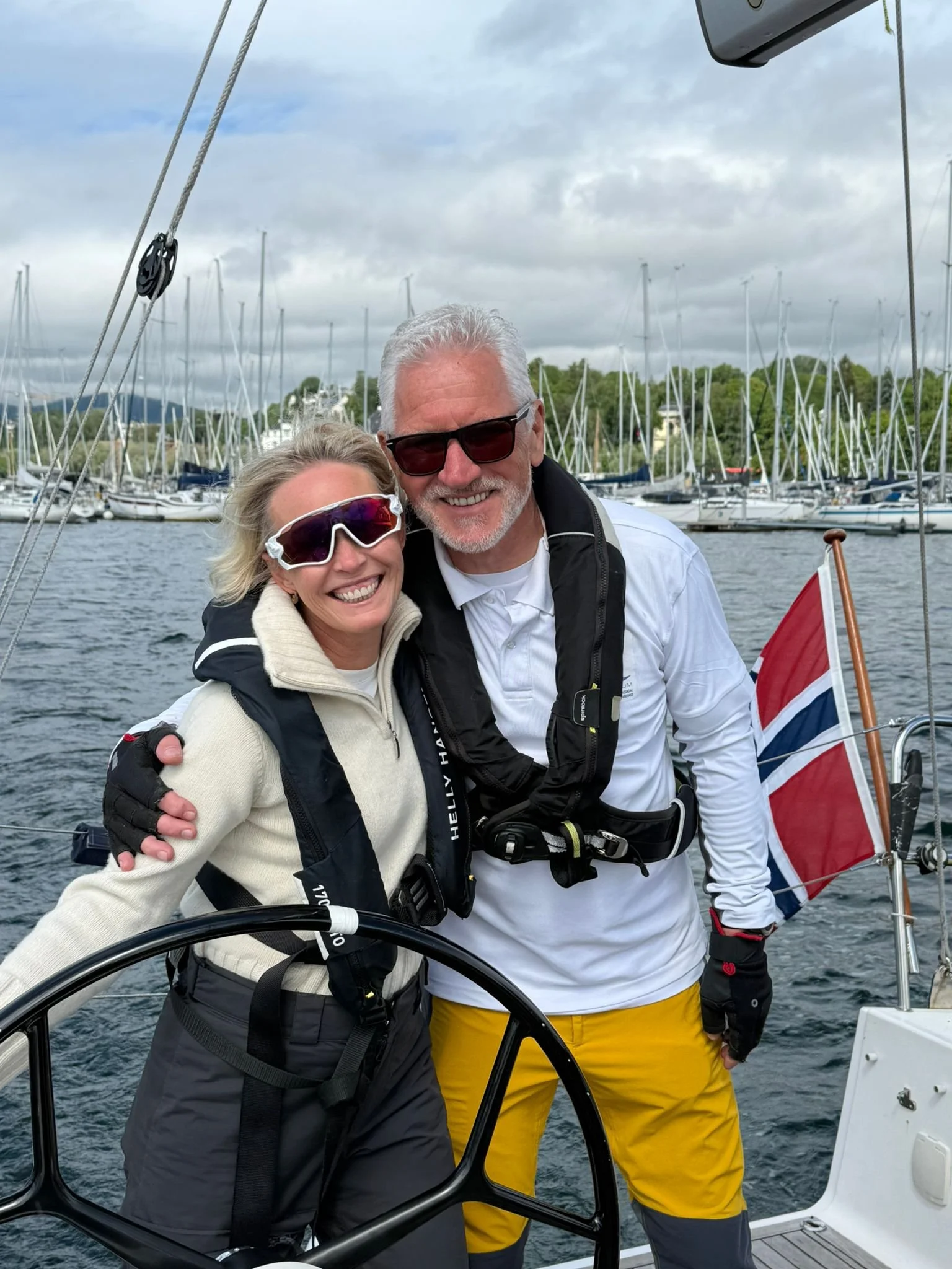 Two smiling adults, a woman and a man, stand on a boat with sailboats and marina in the background. They are wearing sunglasses and jackets, enjoying a cloudy day on the water.