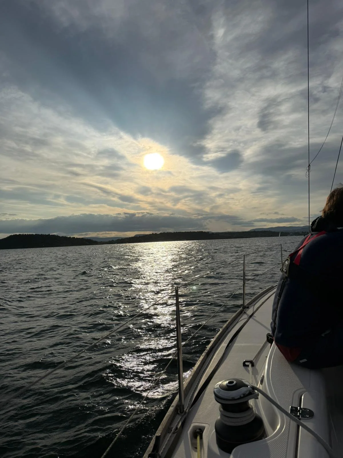 View from a sailboat on the water during sunset, with a person on the right side, the boat's rigging and equipment visible, calm water reflecting sunlight, and distant land with trees and hills under a partly cloudy sky.