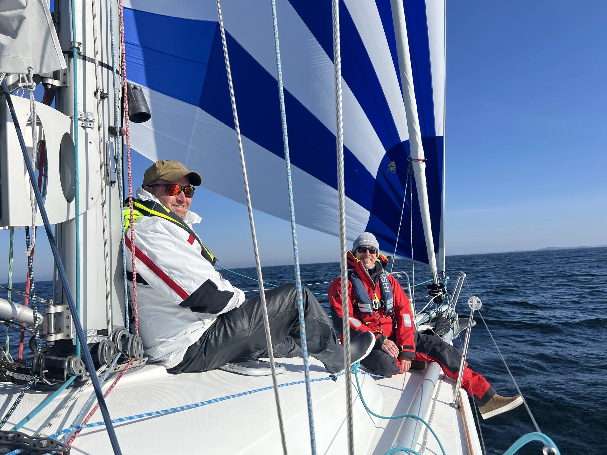 Two men are sitting on a boat in the ocean, smiling and looking towards the camera, with the blue sea and clear sky in the background. One man is wearing a yellow cap, sunglasses, and a white jacket, while the other is wearing black sunglasses, red jacket, and a black hat with white. The sailboat has a large blue and white sail, various ropes, and equipment visible.