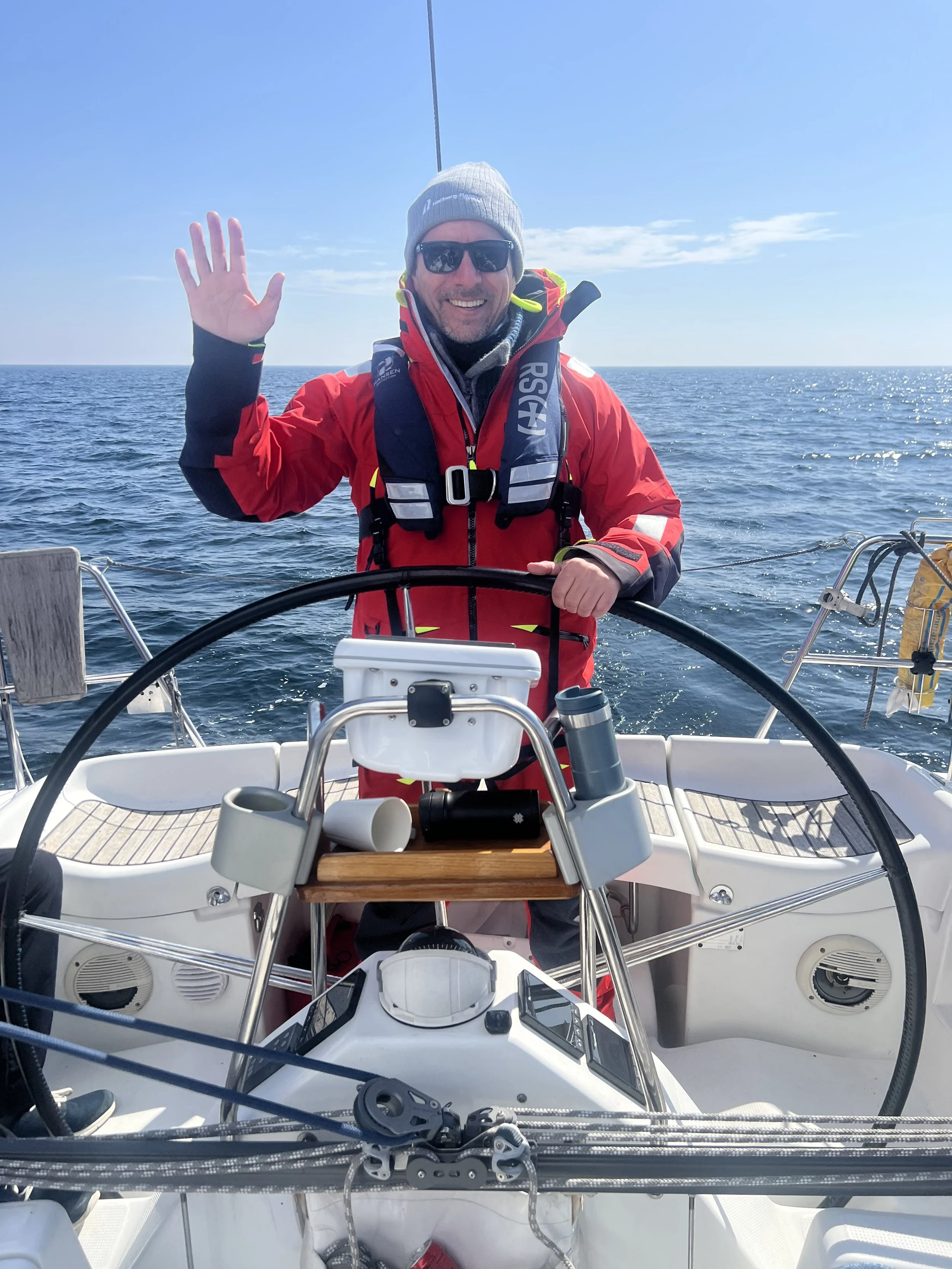 A man in a red jacket, gray beanie, and sunglasses smiling and waving while steering a sailboat on a calm ocean with clear blue sky.