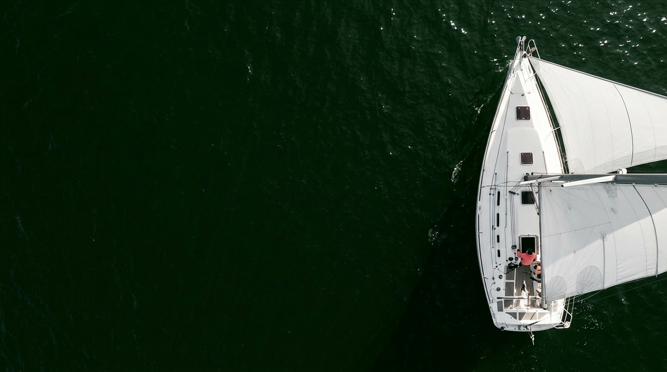 Top-down aerial view of a white sailing boat with two people on deck, floating in dark green water.