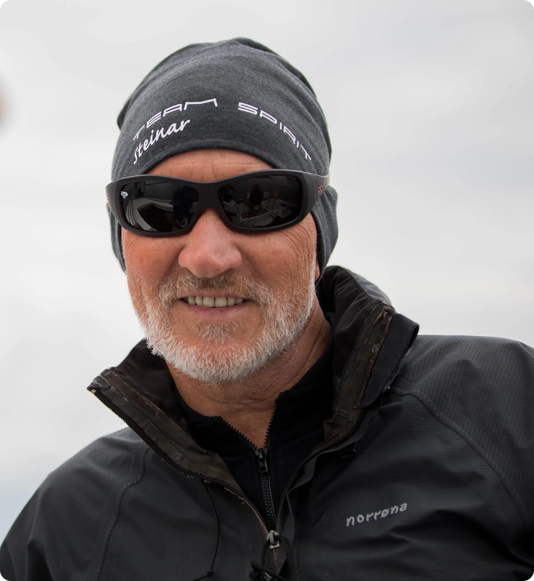 A person with gray hair and beard wearing a gray beanie that reads 'STEAM SPORT' and 'Steinar' and black sunglasses, smiling outdoors against a light, cloudy sky background.