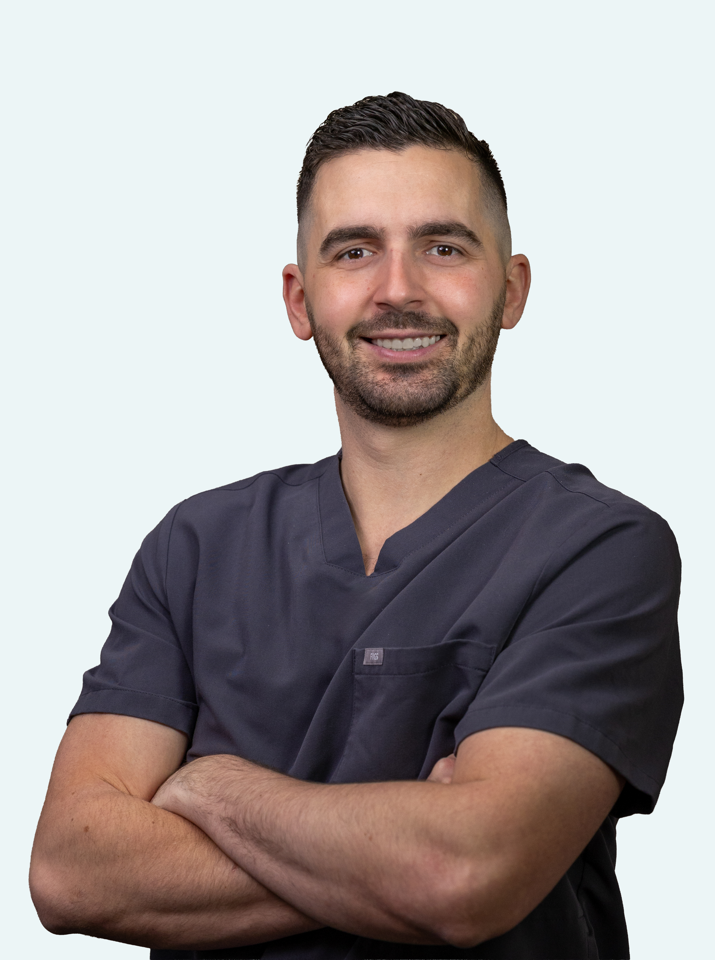 Portrait of a smiling male healthcare professional in dark scrubs with arms crossed, standing against a light background.