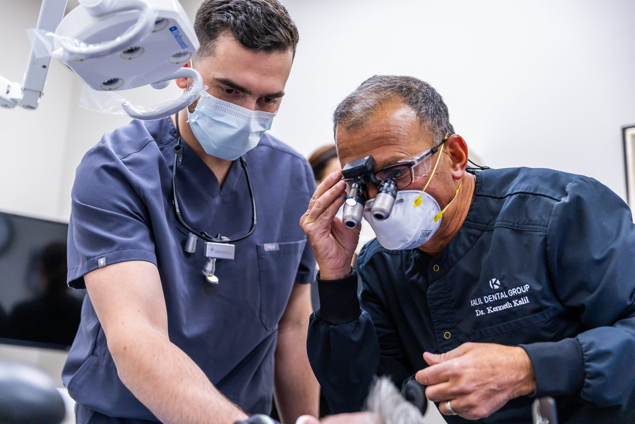Two dentists wearing protective masks and scrubs examining a patient with dental equipment, one using head-mounted magnifying glasses.