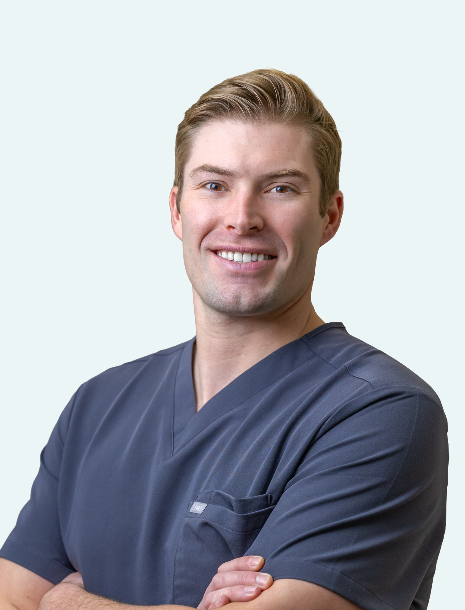 A smiling male healthcare professional with short blonde hair and blue eyes, wearing navy scrubs, standing with arms crossed against a light background.