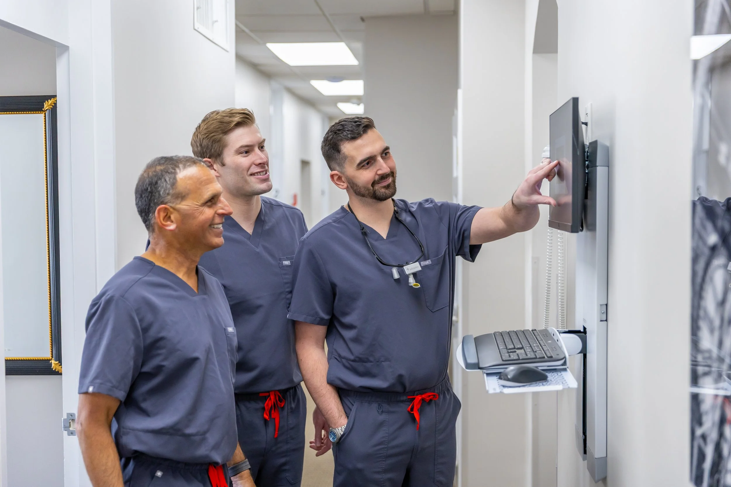 Three male medical professionals in navy scrubs standing together in a hospital hallway, looking at a mounted monitor with one of them pointing at the screen.