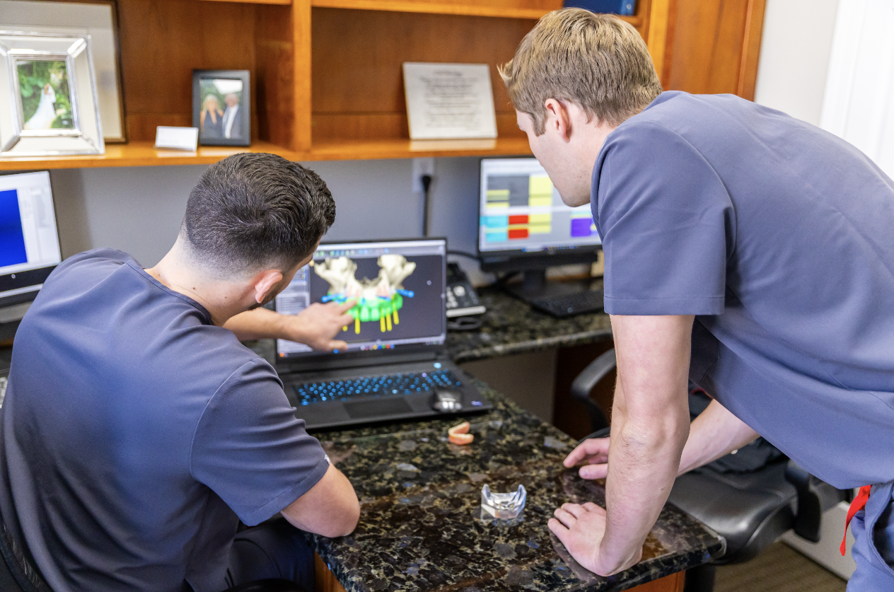 Two men in gray scrubs working together at a desk, one pointing at a computer screen displaying 3D dental models, in an office with multiple monitors and framed photographs.