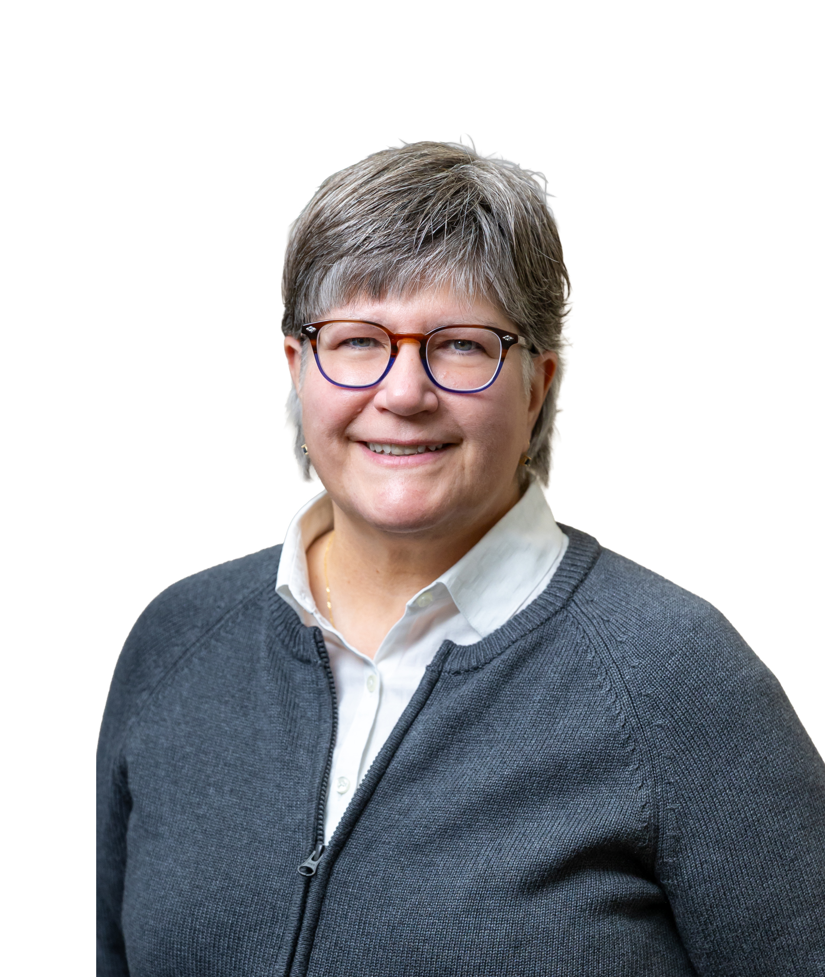 A woman with short gray hair and glasses, smiling, wearing a white collared shirt and a dark gray zip-up sweater against a white background.