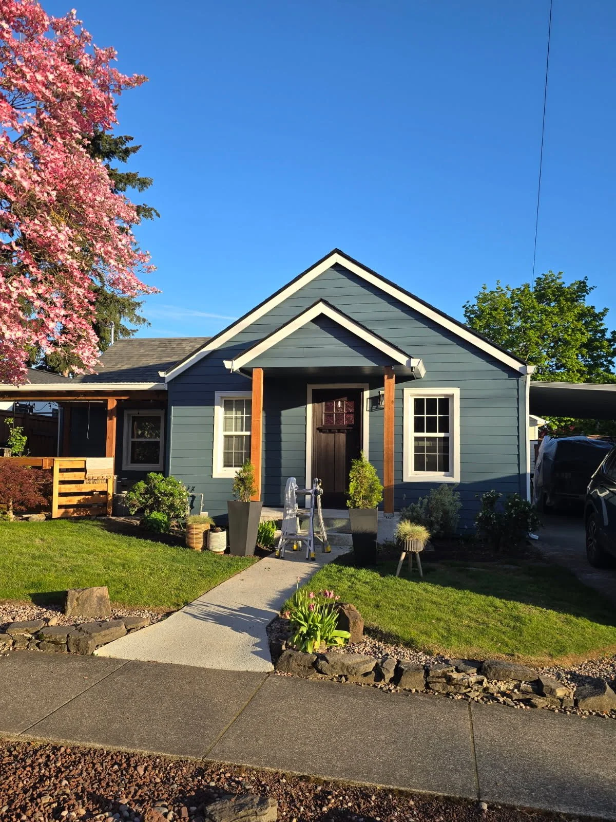Front view of a blue house with white trim, a small porch with potted plants, a walkway, green lawn, and pink flowering tree to the left under a clear blue sky.