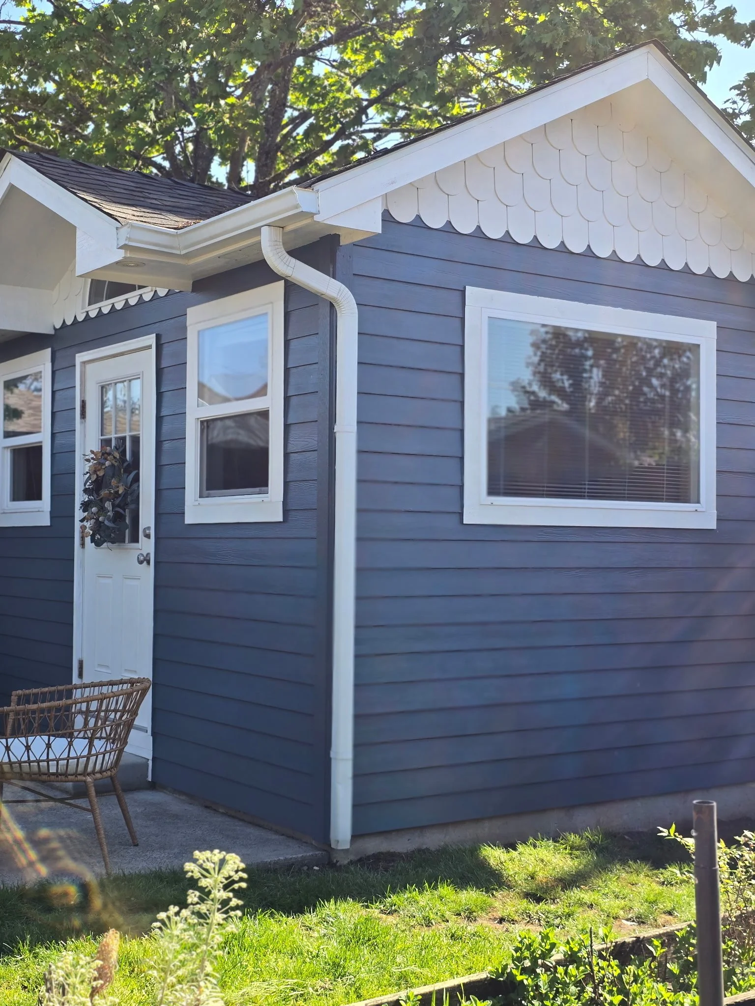 A blue house with white trim, on a grassy lawn, with a wicker chair outside and a small garden patch in the foreground.