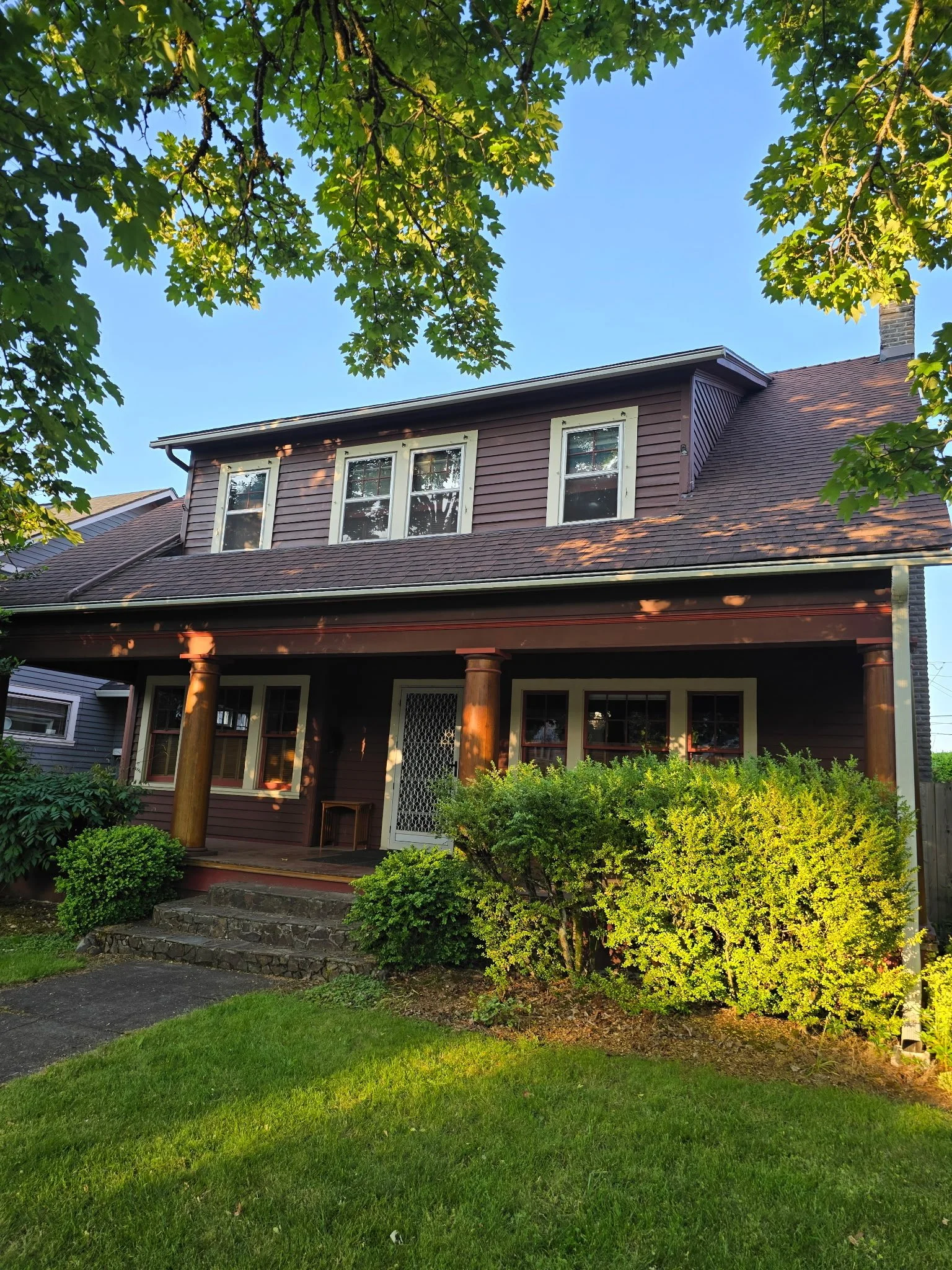 A two-story house with a porch, brown siding, white-framed windows, and lush green bushes in the front yard, under a canopy of leafy trees with blue sky in the background. budger=t friendly paint project