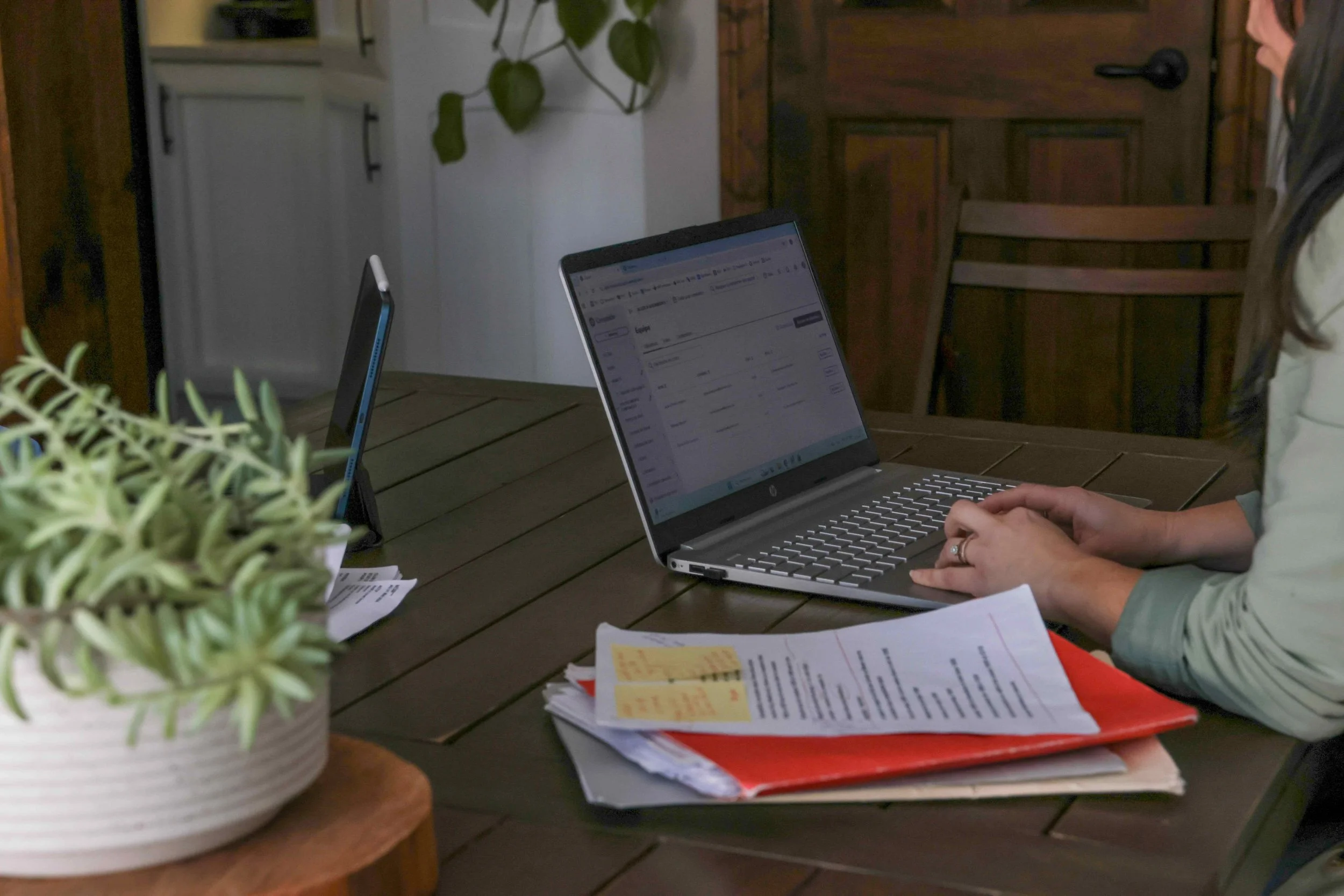 Une personne utilise un ordinateur portable sur une table en bois, au milieu de documents, avec des plantes en pot à gauche.
