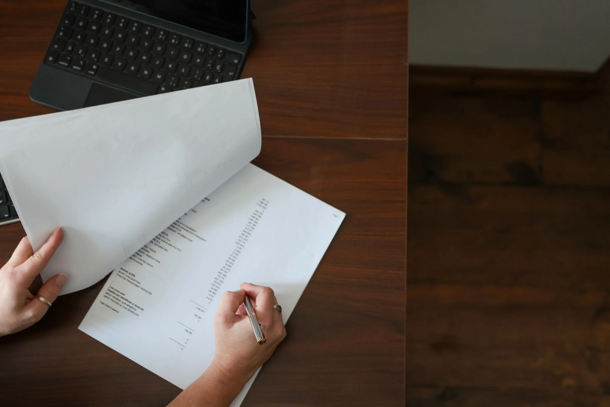 Une personne tient une feuille de papier en train de signer un document financier ou une facture sur un bureau en bois, avec un clavier d'ordinateur portable en haut à gauche.