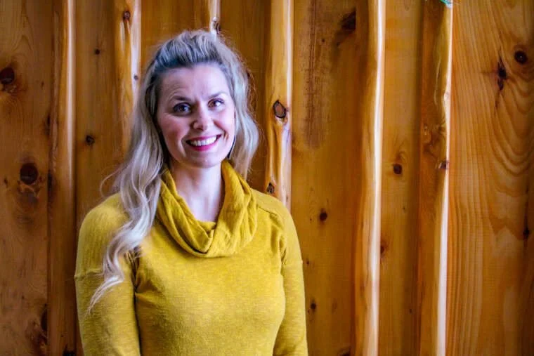 A woman with long blonde hair, smiling, wearing a yellow sweater, standing in front of a wooden wall.