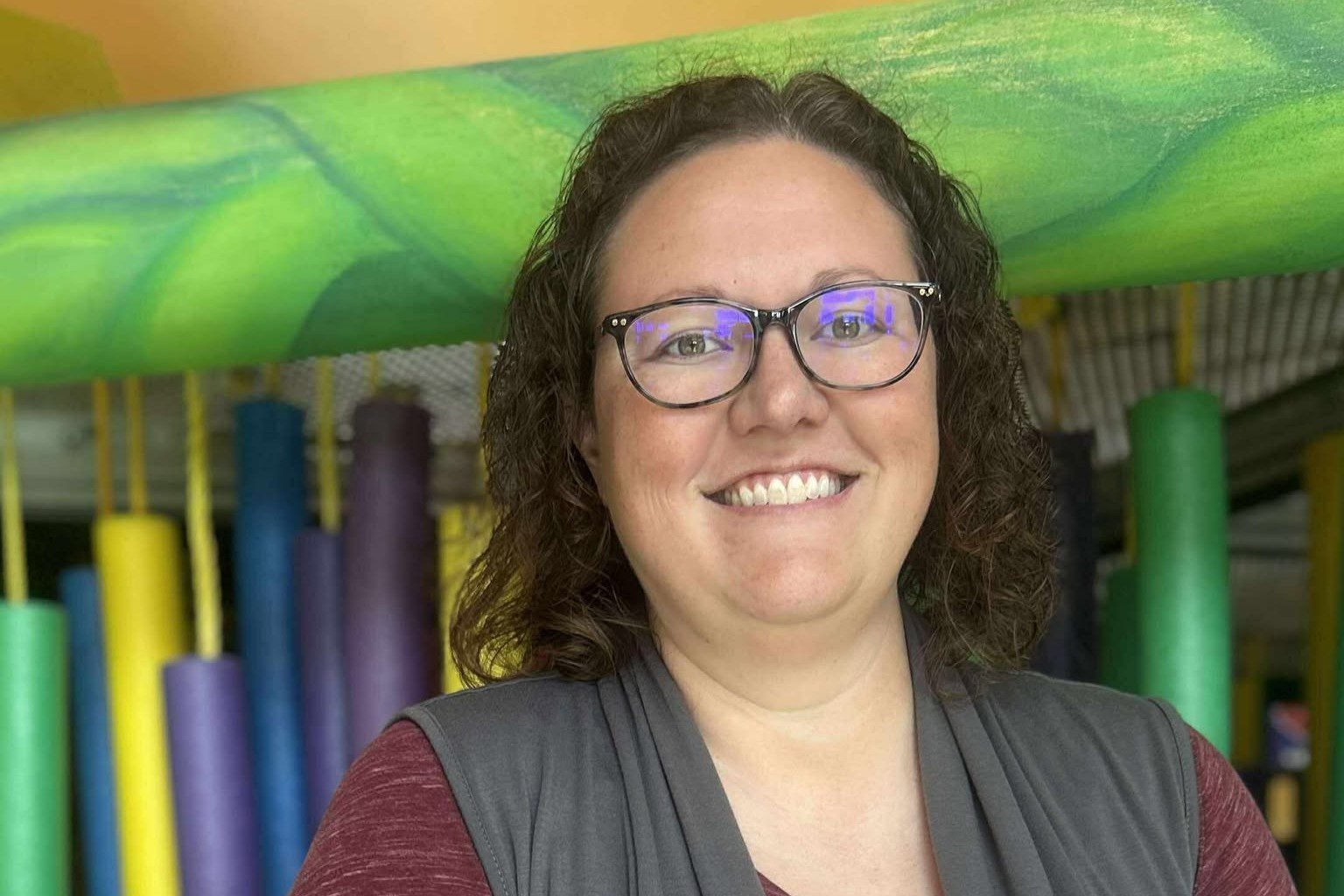 Smiling woman with glasses in indoor play area with colorful foam tubes and structures in the background.