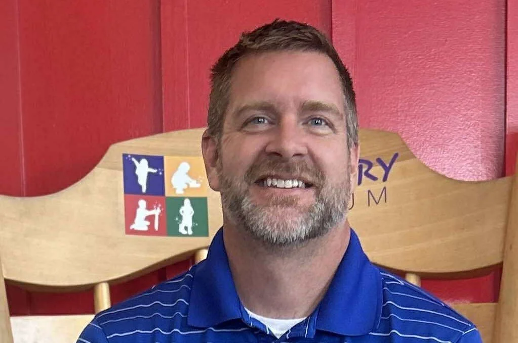A man with short brown hair and a beard, smiling, wearing a blue polo shirt, sitting in front of a wooden chair with colorful icons and text on it.